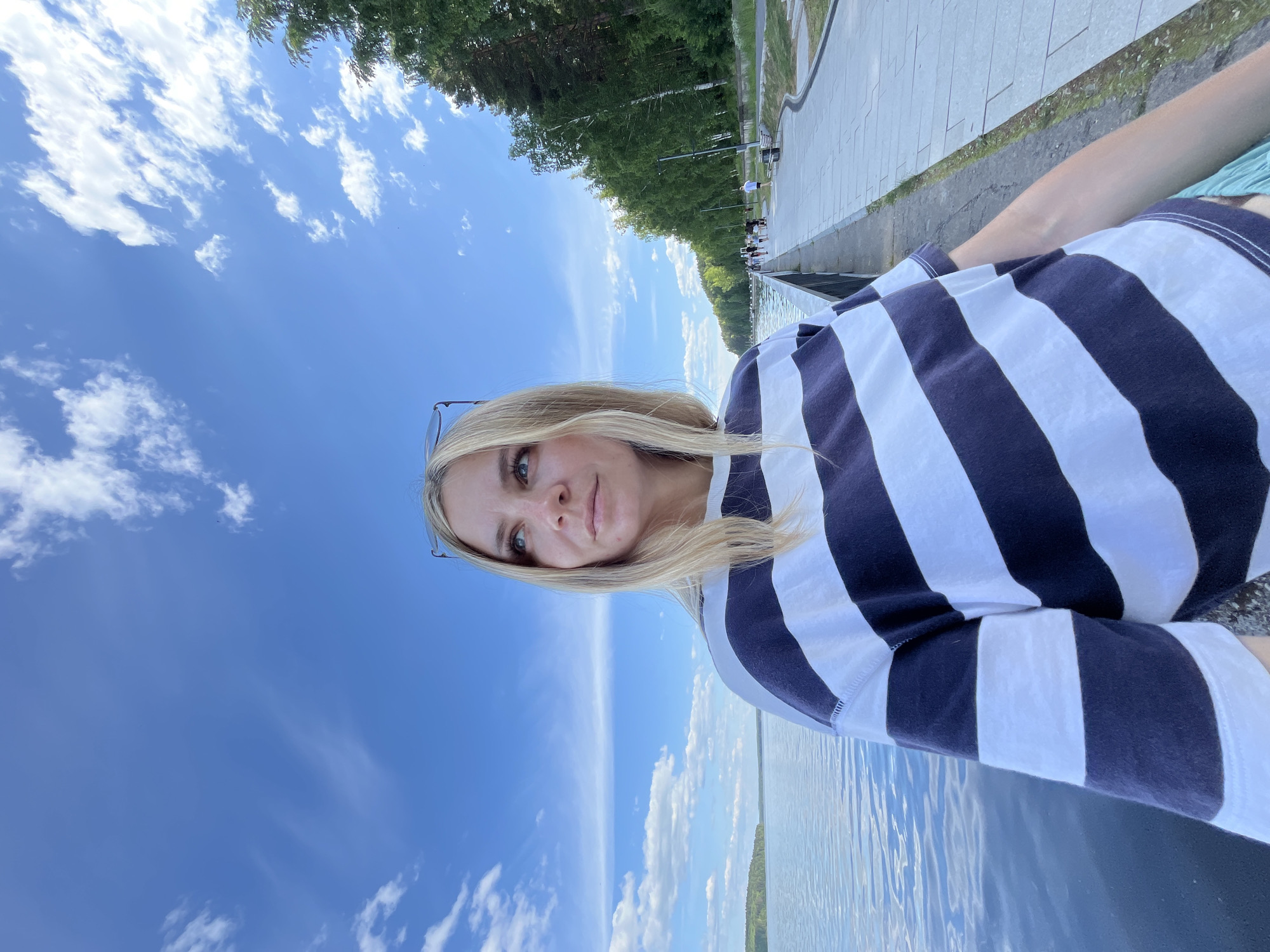 A fair-skinned individual, appearing to be an adult female with long blonde hair, is prominently featured slightly left of the image center. She is wearing a loose-fitting short-sleeved t-shirt with horizontal alternating navy blue and white stripes. A pair of eyeglasses rests on top of her head. Her head is tilted slightly upwards, and her gaze is directed towards the upper left quadrant of the frame.
The background on the left consists of a wide body of calm, dark blue water, extending to a distant forested landmass on the horizon under a clear blue sky with scattered white clouds. The background on the right features a paved promenade or walkway running alongside the water, separated by a low barrier or railing. Several distant figures are visible walking on the promenade. Behind the promenade, a dense line of tall green deciduous trees forms a natural backdrop. Streetlights and benches are also visible along the walkway. This scene depicts an embankment or riverside park along a large body of water, consistent with the Votkinsk Reservoir in Chaikovsky, Russia.