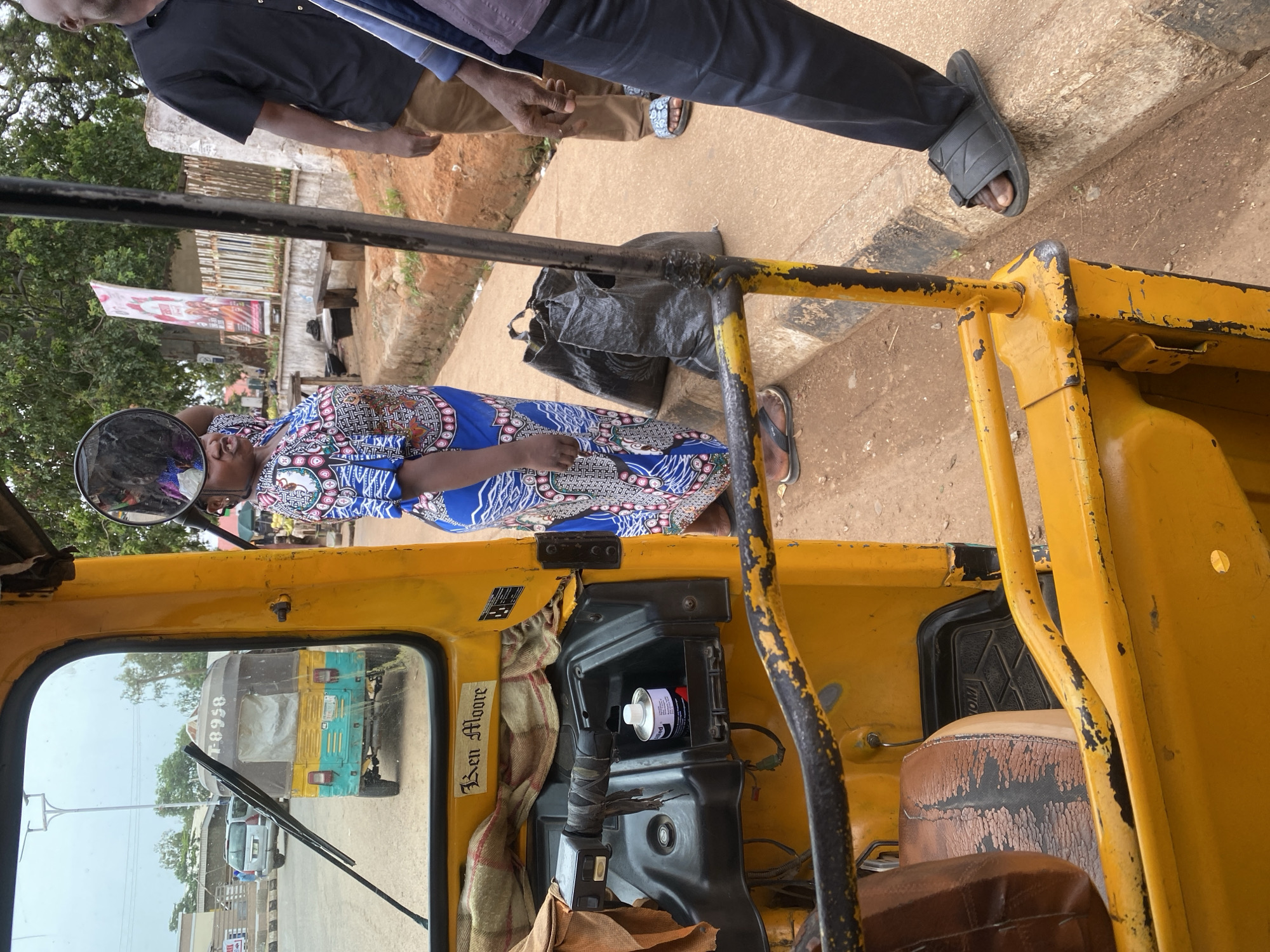 An outdoor, medium shot captures a yellow auto-rickshaw, its driver's side interior visible, from a slightly low angle. The auto-rickshaw is parked on a dirt road beside a curb, indicating it is in a street environment in the city of Jos, Nigeria. In the background, a green and yellow auto-rickshaw with registration number T0958 is parked. The main subject, a woman, stands to the right of the yellow auto-rickshaw, looking towards the left. She is wearing a blue and white patterned dress and holding a round mirror. To her right, a man in a black t-shirt and brown pants stands facing away from the camera, and further to the right, another person in a dark blue top and pants is partially visible. Two large black plastic bags are placed on the ground between the woman and the curb. The environment appears to be a typical street scene with trees and buildings in the background. The auto-rickshaw's interior shows a worn brown seat and a compartment with a can of lubricant.