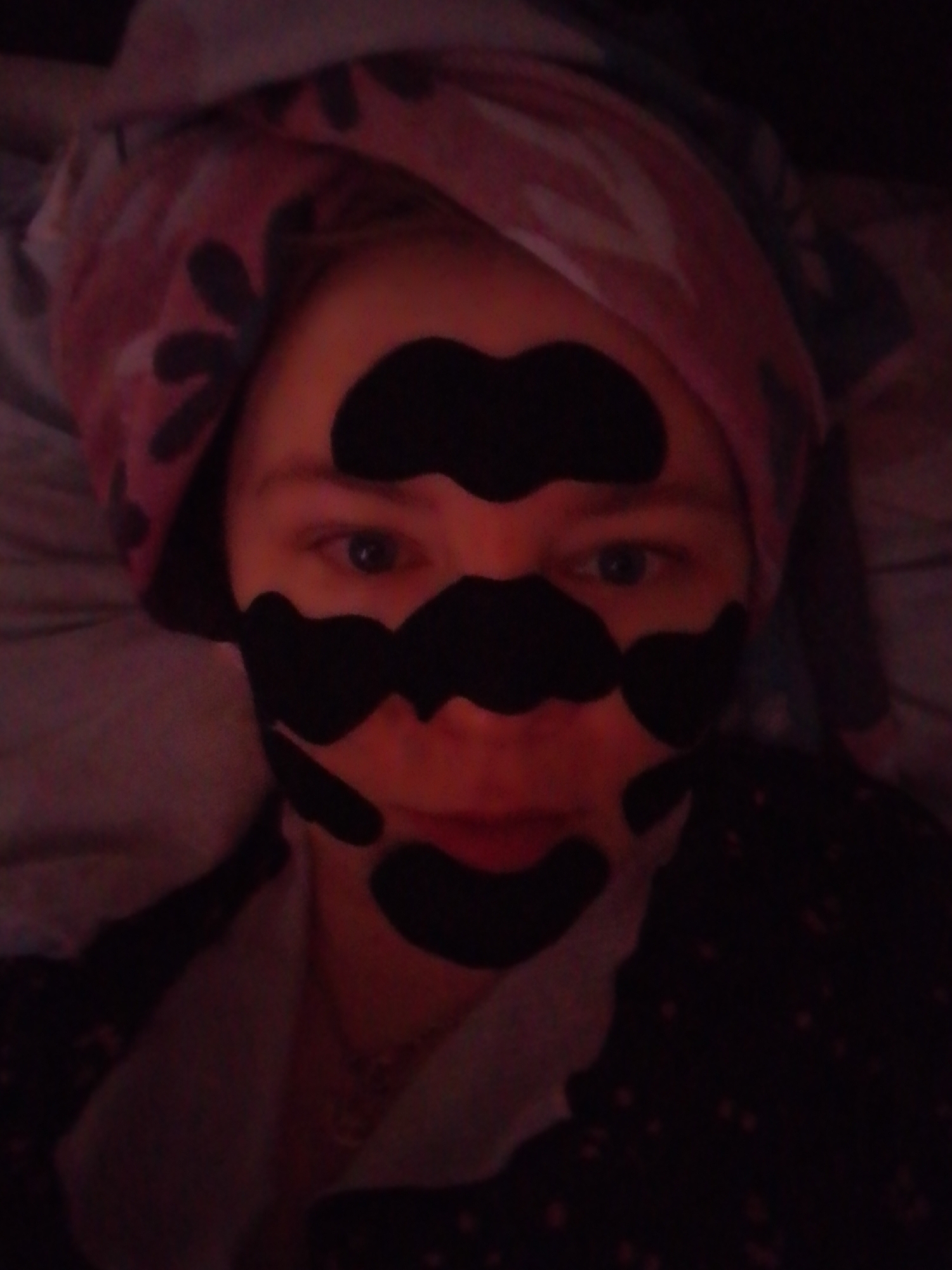 A woman is lying down, wearing a pink and black floral towel wrapped around her head, and multiple black adhesive strips on her face. The background appears to be bedding and possibly pajamas. The lighting is dim and reddish, suggesting an indoor setting with warm-toned artificial light.
There is no information in the image to indicate the location is in Izhevsk, Russia.