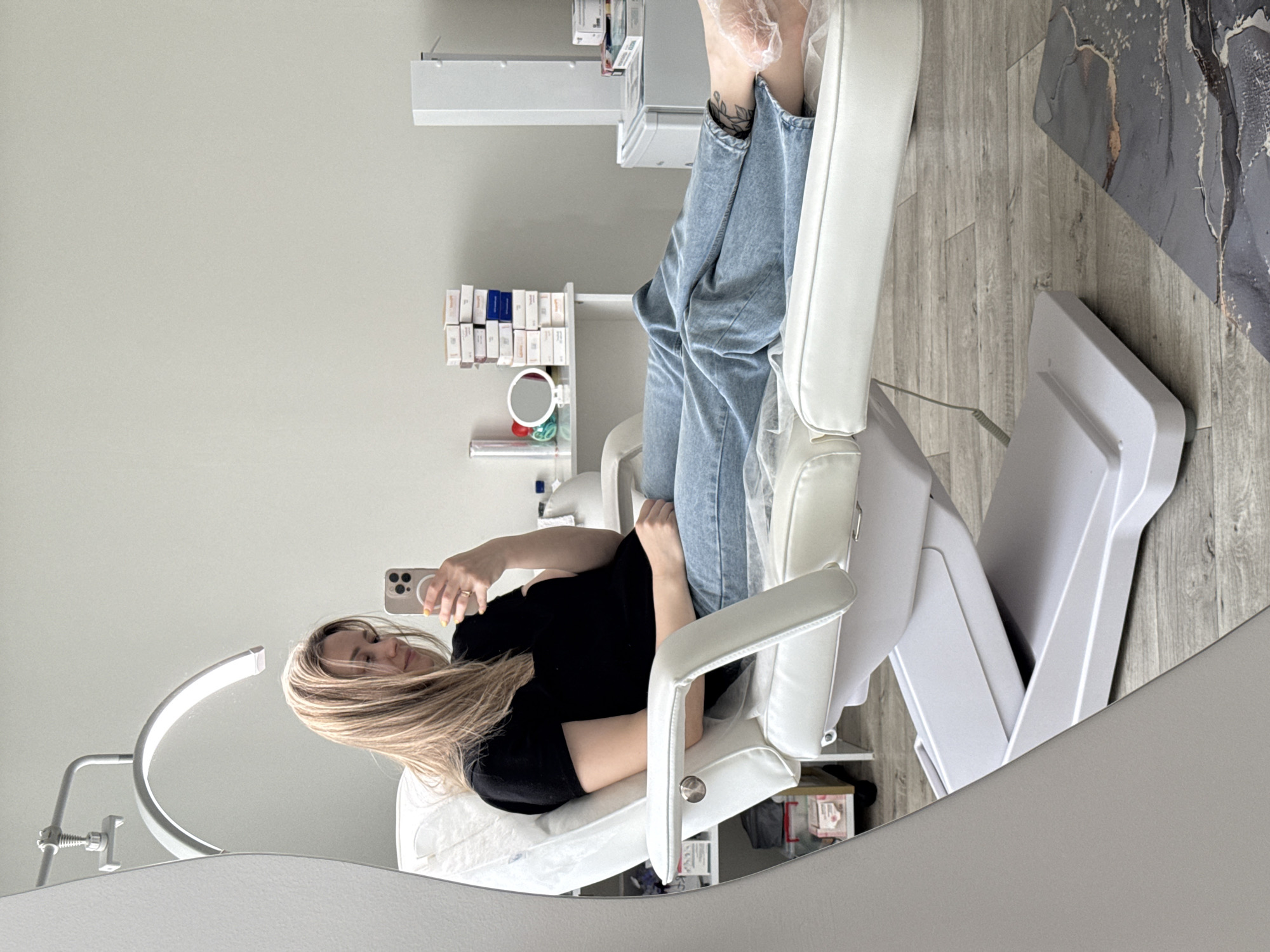 A young woman with long blonde hair, dressed in a black t-shirt and light blue jeans, is captured in a mirror selfie within what appears to be a modern beauty salon or aesthetic clinic in Chaikovsky, Russia. She is seated in a white, adjustable salon chair, holding a smartphone with her right hand, seemingly taking the picture. Her left arm rests across her body.
A notable detail is her left foot, which is covered in a clear plastic bag or protective sleeve, suggesting she is undergoing or has just completed a foot or leg treatment. A tattoo, possibly a leafy or floral design, is visible on her lower left leg near the ankle.
The setting features light grey walls and light wood-look flooring. Behind the woman, a white shelving unit holds numerous small, rectangular white boxes, some marked with "P" and "LUMION", indicating they are likely cosmetic products or supplies. A small round mirror also sits on one of these shelves. To the left of the woman, a distinctive curved LED lamp, commonly used for magnifying and illuminating work in beauty treatments, is visible. Other white equipment and furniture are present throughout the room, reinforcing the sterile and professional atmosphere of a beauty or wellness establishment. The floor also has a dark grey, abstractly patterned rug or mat.