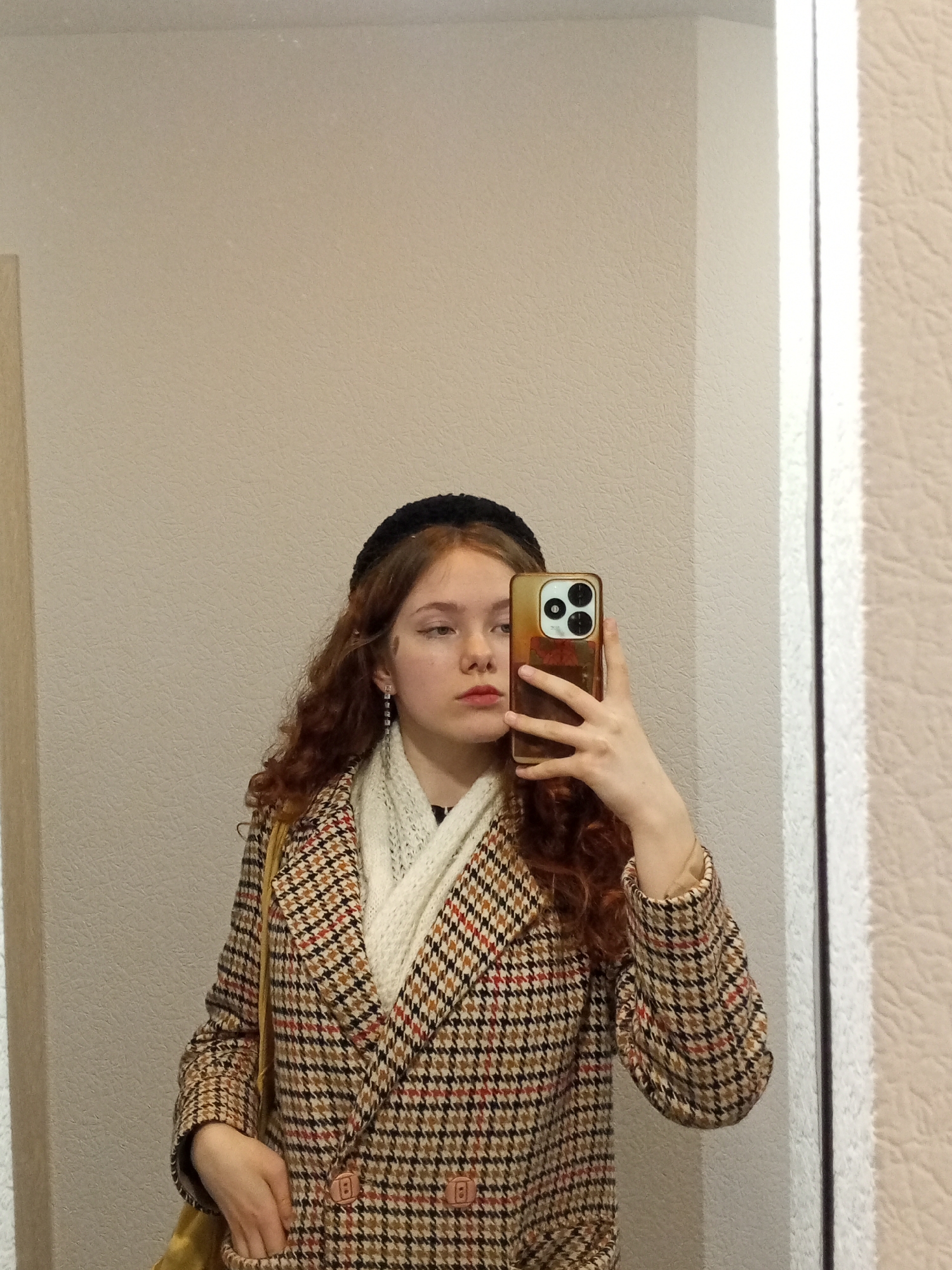 A young, light-skinned woman with reddish-brown curly hair and a black headband is taking a selfie in a mirror. She is wearing a brown and black houndstooth patterned coat over a white scarf or shawl. She is holding a gold-colored smartphone with three camera lenses visible. Part of a yellow purse or bag is visible over her shoulder. The mirror frame is white. The walls are painted a light beige color. The exact location within Chaikovsky, Russia, is not determinable from the image.