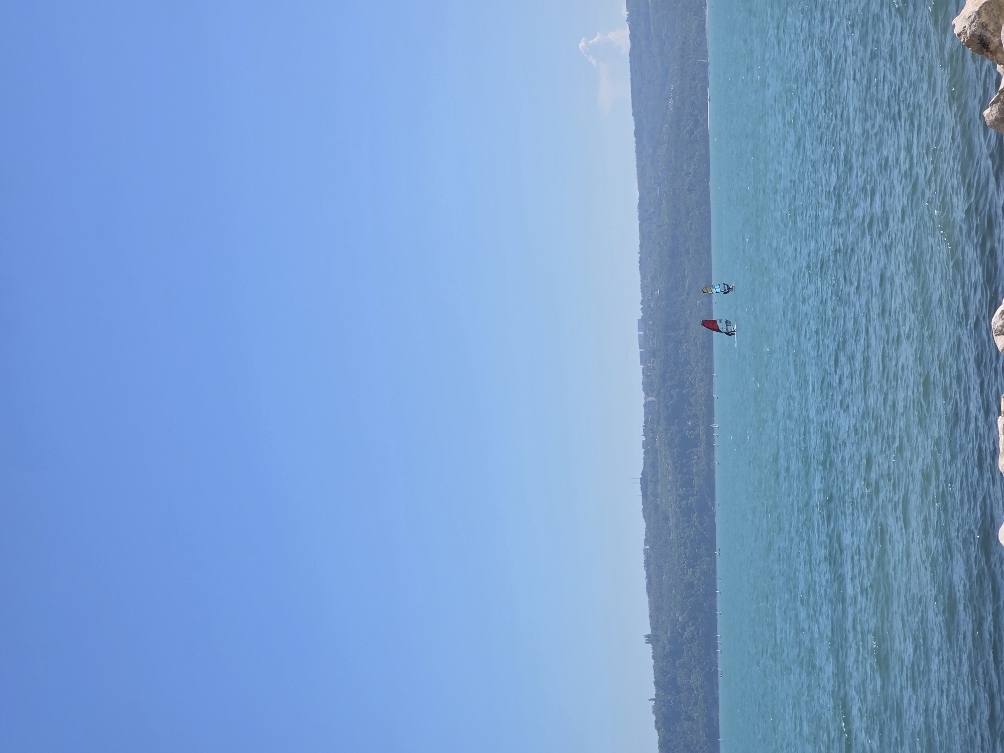 The image depicts a coastal scene, likely a lake, with a bright blue sky occupying the left portion of the frame. The right side shows a body of water with gentle waves, and the coastline is visible in the middle. On the water, there are two windsurfers with sails of different colors (red and blue). The coastline appears to be vegetated with trees. Some rocks are present on the right edge of the frame. The location is near Varna, Bulgaria, as the landscape is reminiscent of the coastline and lakes in the area.