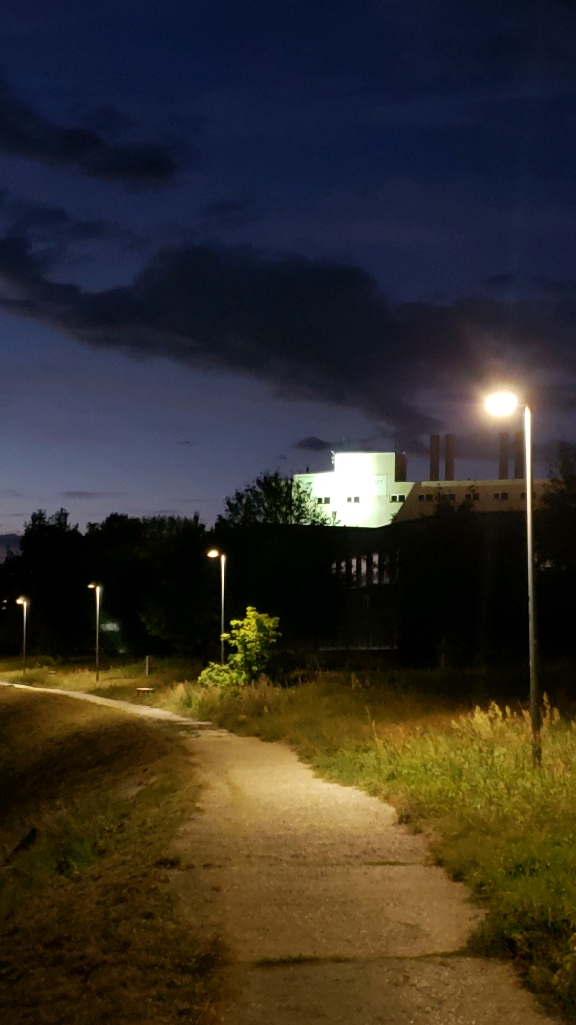 The image depicts an outdoor scene at dusk or night. A paved pathway curves from the foreground into the midground, illuminated by artificial lights from street lamps. The path is bordered by grass and vegetation. In the background, a large, white, illuminated building with multiple windows and tall chimneys is visible, surrounded by trees and more vegetation. Several street lamps are positioned along the pathway and in the surrounding area. The sky above is dark blue with scattered clouds. The overall atmosphere is one of a quiet, possibly industrial or institutional area at night, with artificial lighting providing visibility. The location is not specified in the image itself.