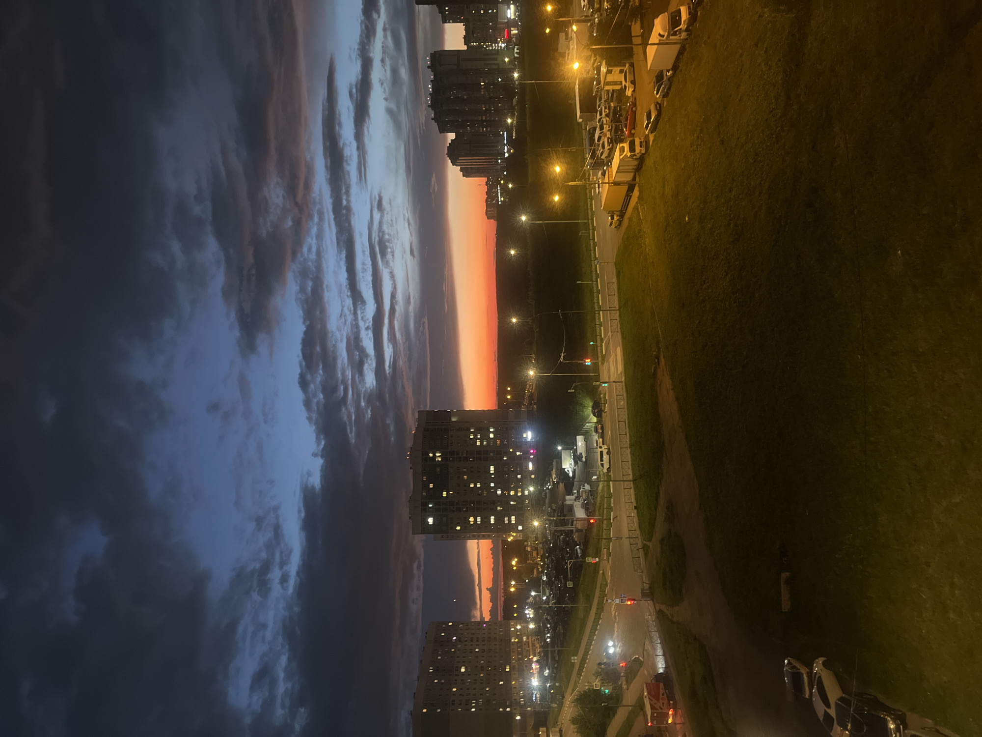 This is a high-angle, long shot photograph taken at dusk in the city of Domodedovo, Russia. The sky is a dramatic mix of dark, brooding clouds and a vibrant orange and blue sunset. In the foreground, a grassy hill slopes down towards a road and a parking lot. Several cars and trucks are parked in the lot, and streetlights illuminate the scene.
Dominating the mid-ground are two large apartment buildings, their windows illuminated, suggesting residents are home. In the distance, more city buildings are visible, silhouetted against the colorful sky. A few cars are on the road, their headlights creating streaks of light. The overall impression is one of an urban landscape transitioning from day to night under a striking sky.