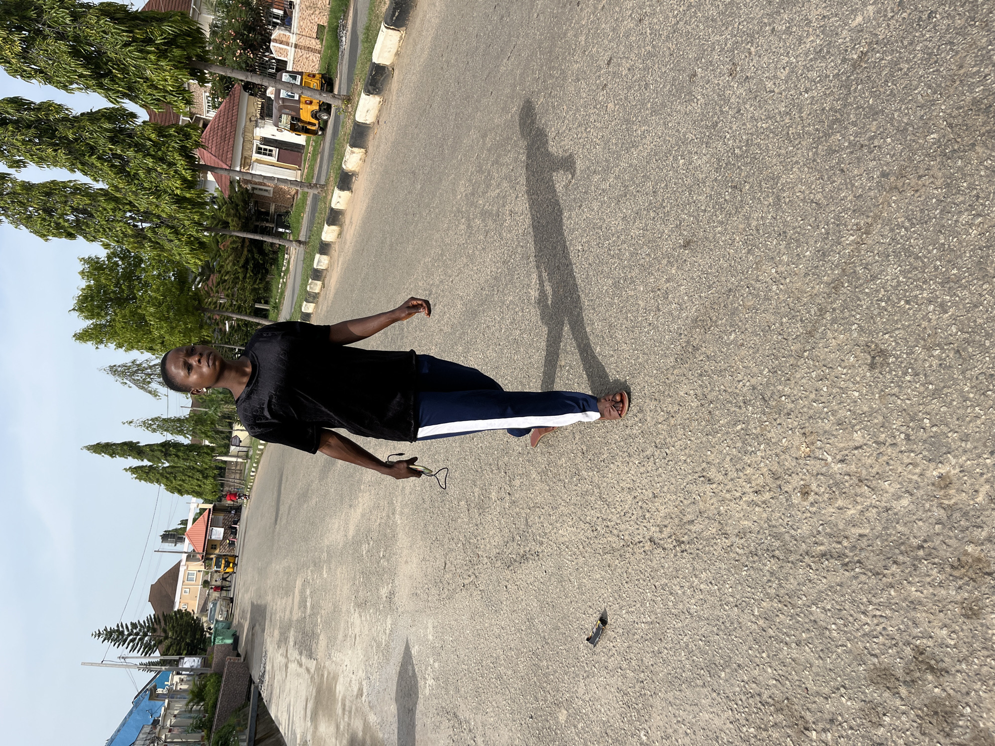 A medium shot of a young man standing on an asphalt road in Abuja, Nigeria. The man is wearing a black T-shirt and blue pants with a white stripe. He is holding a pair of glasses in his right hand. His shadow is cast on the road to his right. The background shows houses, trees, and a yellow vehicle. The road has white and black painted curbs.