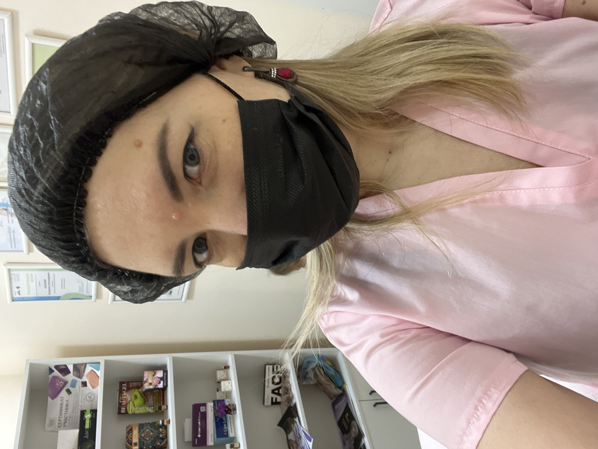 The image shows a woman wearing a pink scrub top, a black disposable mask, and a black disposable hair net. She is wearing a dark-colored earring with a red stone. She has light-colored hair and is wearing eyeliner. She is in what appears to be a salon or medical office. Behind her, there are shelves with various products and certificates on the wall. The overall impression is that of a professional setting, likely a cosmetic or medical spa. There is no visual information to suggest the location is the city of Sochi, Russia.