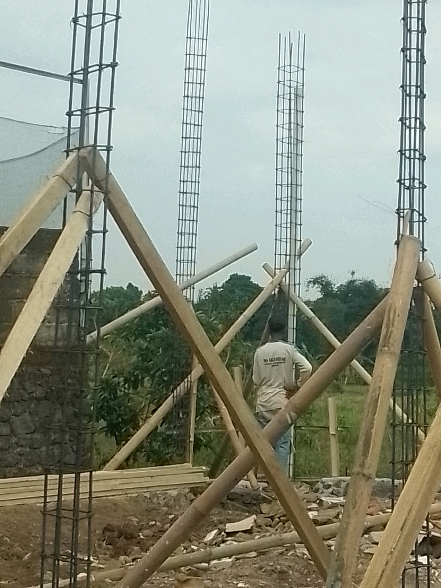 An outdoor construction scene in Jember, Indonesia, shows a man wearing a light-colored long-sleeved shirt and blue pants standing with his hands on his hips, facing away from the camera. He is positioned between scaffolding made of bamboo and wooden beams. In the background, unfinished concrete pillars are visible, reinforced with rebar. The area is cluttered with construction debris and piles of lumber. Lush green trees form the backdrop, suggesting a rural or semi-rural setting. The sky is overcast.