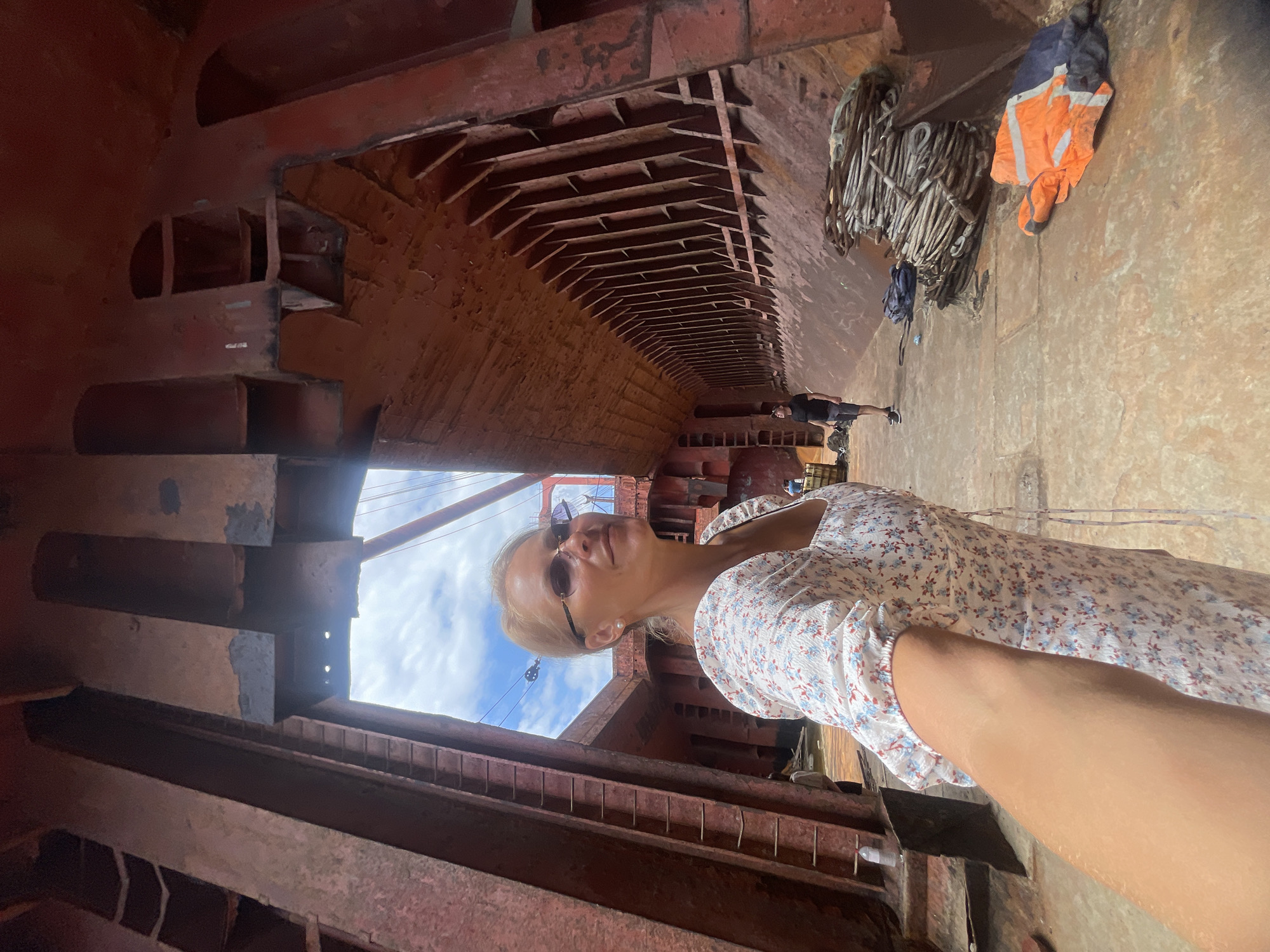 In the dry dock of the city of Kabardinka, Russia, a woman with blonde hair and sunglasses is taking a selfie. She is wearing a white dress with a floral print. The background shows the interior of a ship's hull, with large metal structures and scaffolding. Two other individuals are visible in the distance, one wearing a black t-shirt and shorts, and another partially obscured figure. Various equipment, including ropes and an orange safety vest, are scattered on the floor of the dry dock. The sky is visible above, with clouds and some rigging.