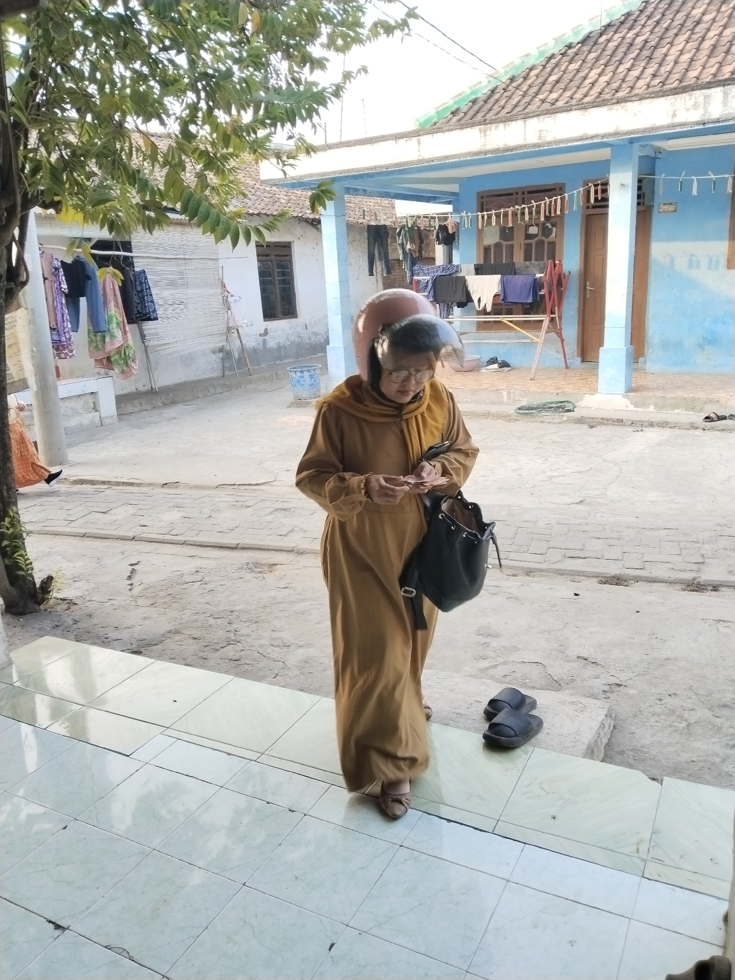 The image depicts a woman wearing a tan robe and a pink helmet, walking outdoors in what appears to be a residential area in Serang, Indonesia. She is holding a small object in her hands and carrying a black purse on her shoulder. The environment features a blue house with a tiled roof, laundry hanging on clotheslines, and paved ground. A pair of black sandals are placed on the ground near the woman. The scene is illuminated by natural daylight, casting shadows and highlighting the textures of the environment.