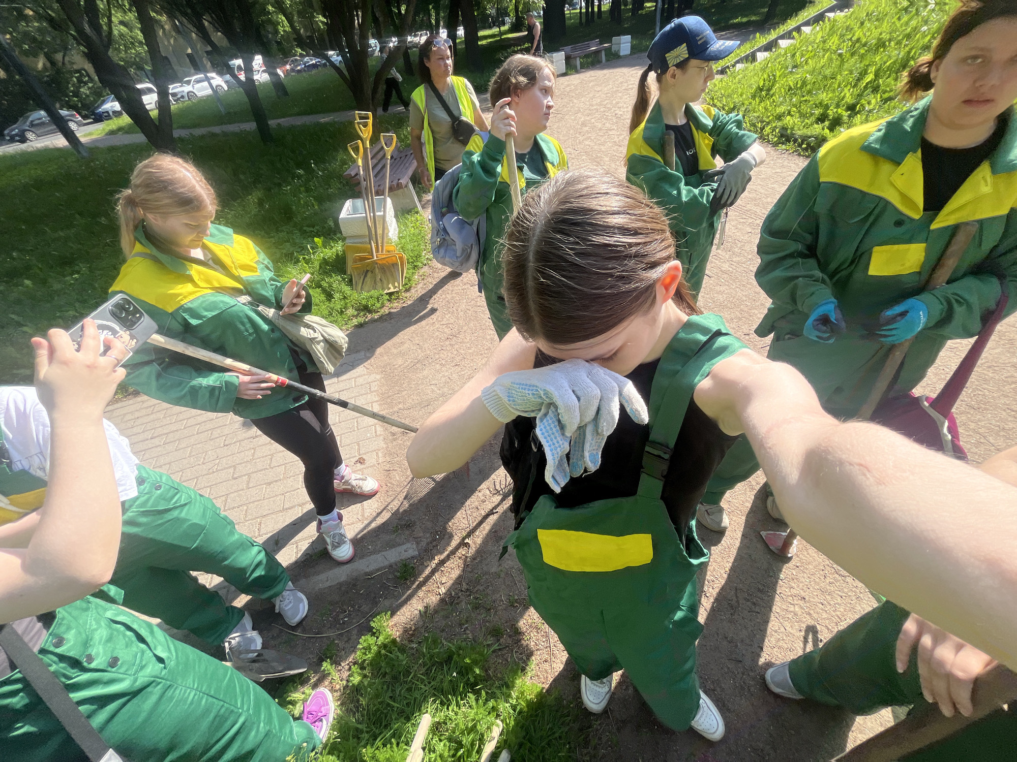 The image depicts a group of people, likely volunteers, engaged in park cleanup or gardening in Saint Petersburg, Russia. They are wearing green and yellow overalls, suggesting a work uniform. Several individuals are holding gardening tools such as rakes and shovels, which are also visible standing in a bucket-like container. The environment appears to be an outdoor park or public space with trees, grass, and a paved walkway. Cars are visible in the background, indicating a street or parking area adjacent to the park. The overall scene suggests a community effort for environmental beautification.