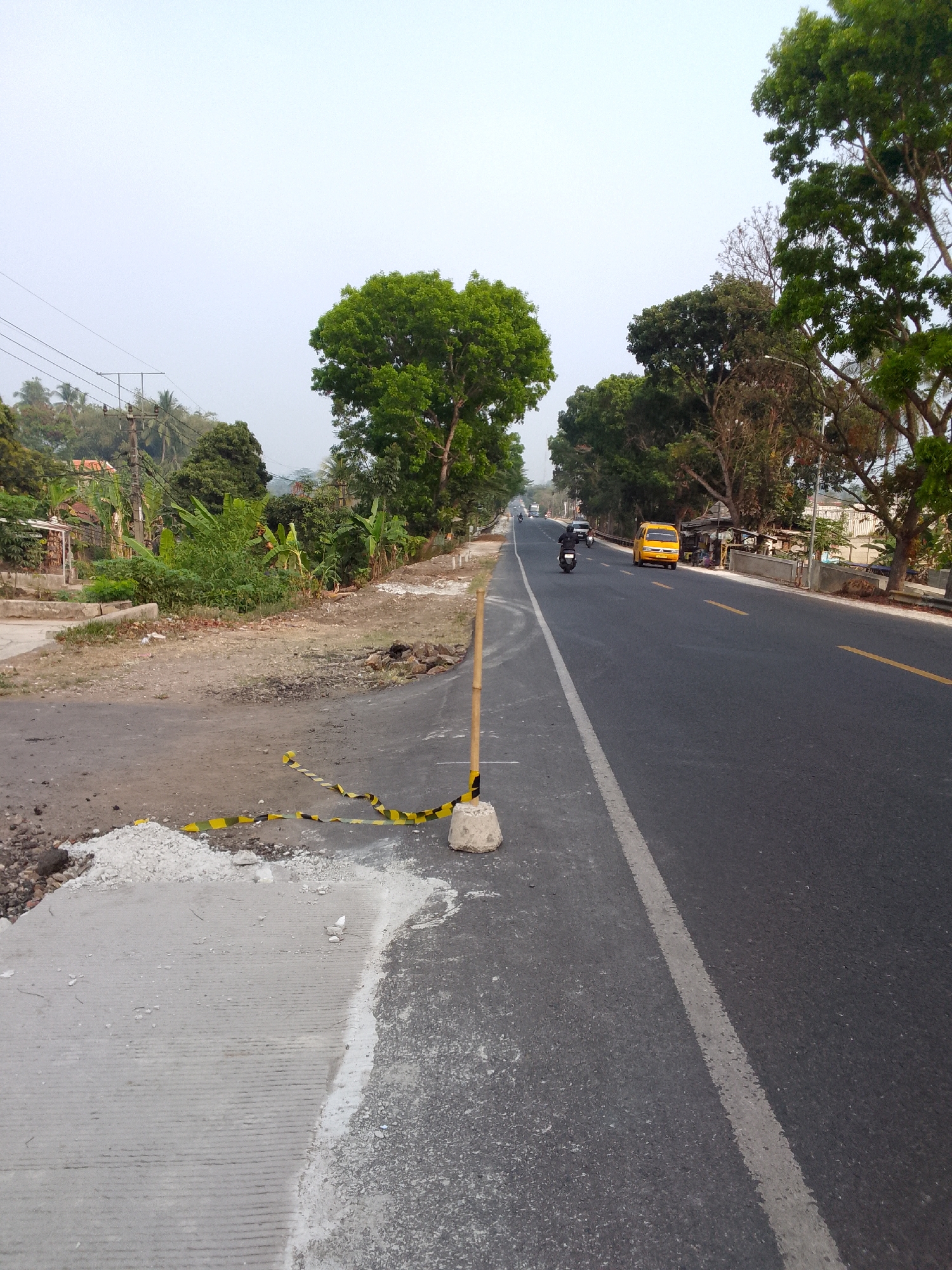 The image captures a scene on a road in Rajamandala Kulon, Indonesia, likely during daytime. The road is paved with asphalt and has a yellow center line and a white lane divider. On the left side of the road, there is a construction area with loose gravel, a patch of concrete with ribbed texture, and yellow and black caution tape. A bamboo pole secured by a concrete base marks the edge of the construction. Beyond the construction, there are trees, utility poles with wires, and some houses. On the right side of the road, the asphalt continues, with a yellow line on the center. Several vehicles are visible: a yellow minivan is parked on the right shoulder, and a motorcycle is in motion on the road. In the background, more trees and a hazy sky complete the scene. The overall impression is of a rural or semi-urban roadside with ongoing infrastructure work.