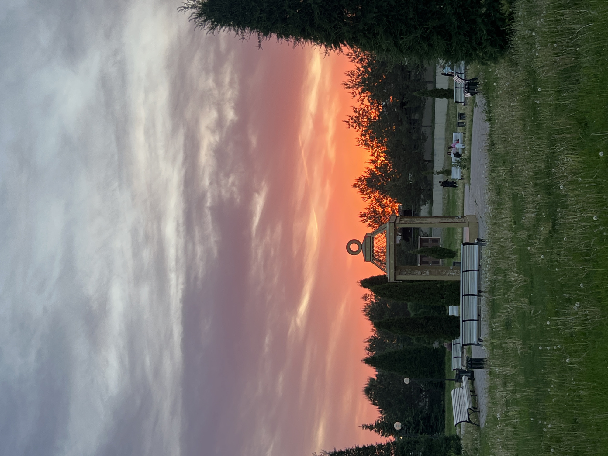 The image captures a scene at Arskoe Cemetery in Kazan, Russia, at twilight. The sky presents a gradient of orange and red hues near the horizon, transitioning to grayish-white above. Silhouetted trees line the horizon. A decorative pergola-like structure with a circular top is visible amidst the trees. Benches are situated along a paved path bordered by grassy areas containing dandelions. Several individuals can be seen on the benches.