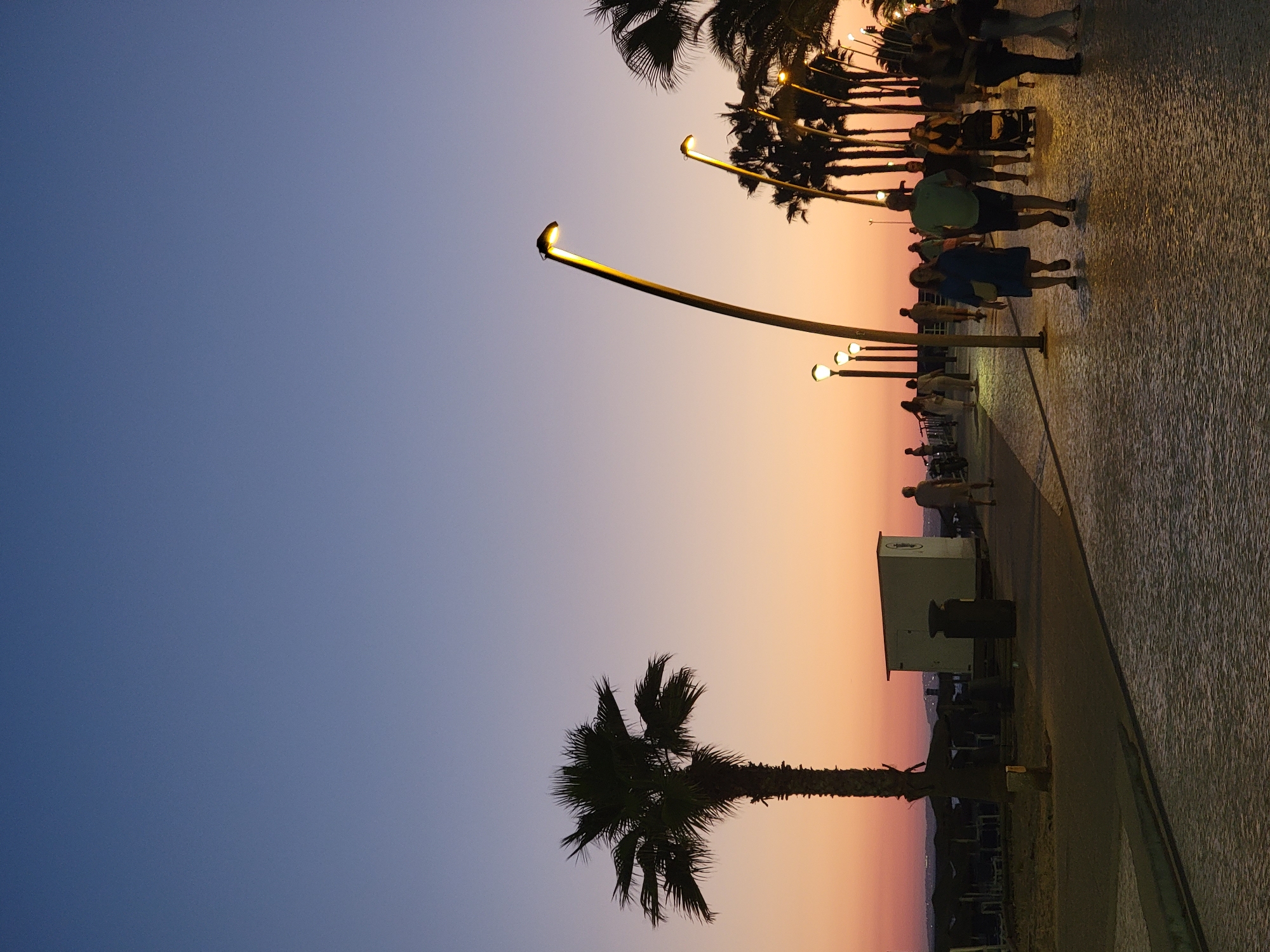 The image depicts a sunset scene on the boardwalk in Quarteira, Portugal. The sky transitions from a deep blue at the top to shades of orange and pink near the horizon. Several palm trees are visible, with one prominent tree silhouetted on the left side of the frame. The boardwalk is paved with cobblestones, and lampposts with warm, yellow lights illuminate the pathway.
A group of people are walking along the boardwalk. In the foreground, a couple is walking towards the camera, with the woman wearing a blue dress and the man in a green t-shirt and dark shorts. Behind them, other individuals and a person pushing a stroller are also visible. To the left of the boardwalk, there is a white structure with a dark trash receptacle. Further in the background, beach umbrellas and chairs are set up, indicating the proximity of the beach. The overall atmosphere suggests a pleasant evening stroll in a coastal city.
