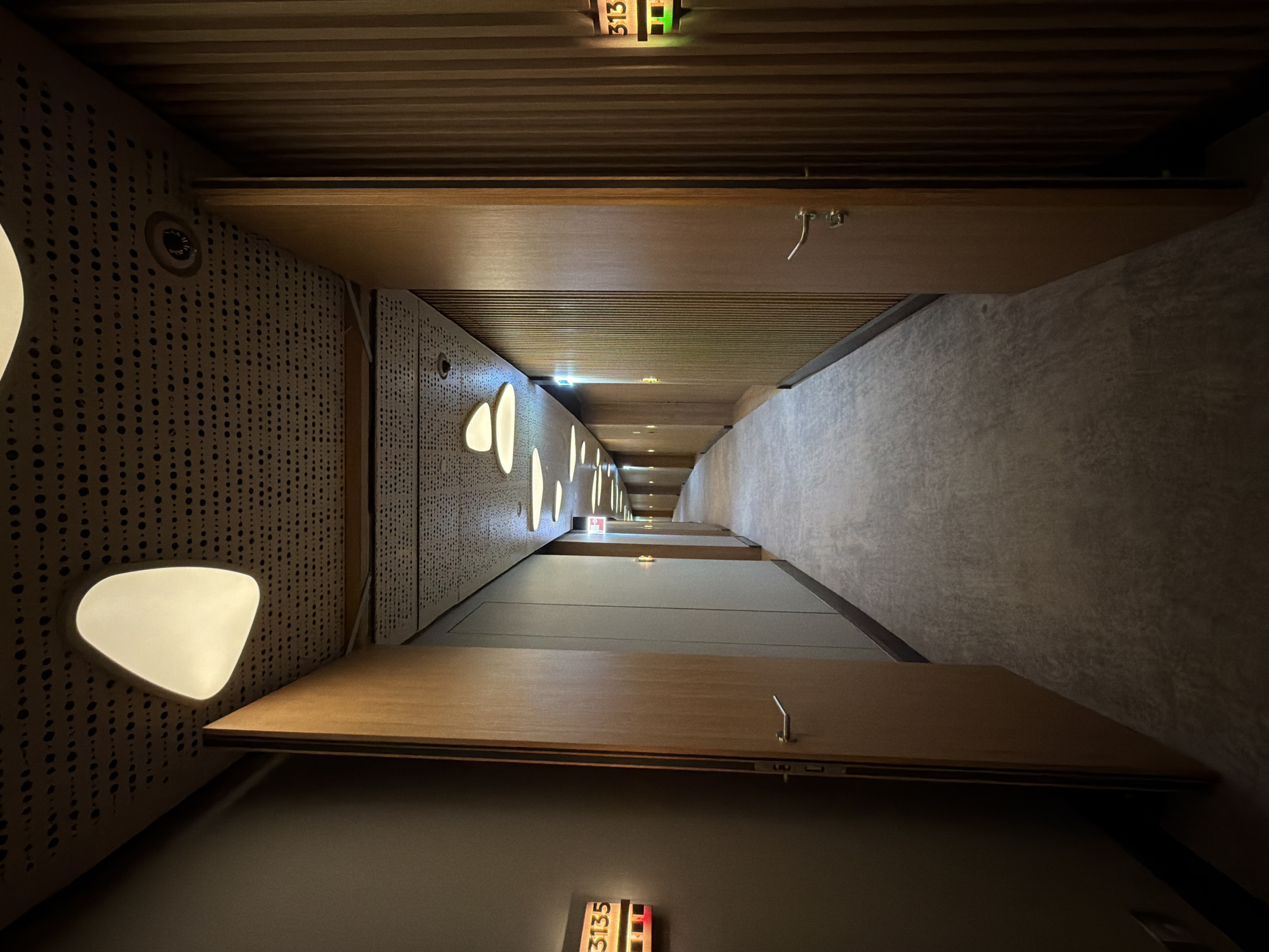 The image shows a hallway in a building, possibly a hotel, with a narrow perspective. The walls are decorated with perforated panels and illuminated with teardrop-shaped lights. The hallway is lined with wooden doors, each displaying a room number. The room numbers visible are "313" and "3135". The ceiling is constructed with parallel wooden planks. The image does not contain any identifiable people or actions. There is no information available to determine the location is in Озёрное, Russia.