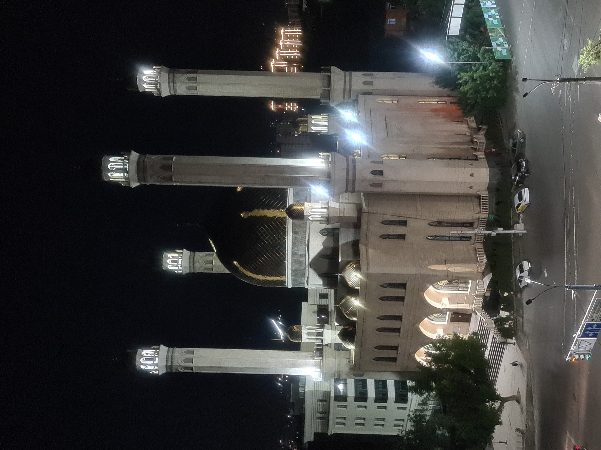 The image shows a large, ornate building with multiple minarets, likely a mosque, at night. The building is illuminated with interior lights, highlighting its architectural details. Several tall, slender minarets with illuminated tops stand prominently. The main structure features arched windows and a large golden dome.
The scene appears to be a city at night, with a street running alongside the building. Cars are visible on the street, and streetlights cast illumination. There are also trees and some buildings in the background. A traffic light is visible, indicating an intersection.
The time of day is clearly night, as the sky is dark and artificial lights are the primary source of illumination. There are no visible people in the frame. The weather appears to be clear, with no indication of rain or fog.
Location cues suggest Almaty, Kazakhstan, though this is not explicitly stated in the image itself. The overall mood is one of quiet grandeur, with the illuminated building standing as a focal point against the dark night sky. There is no visible text in the image.