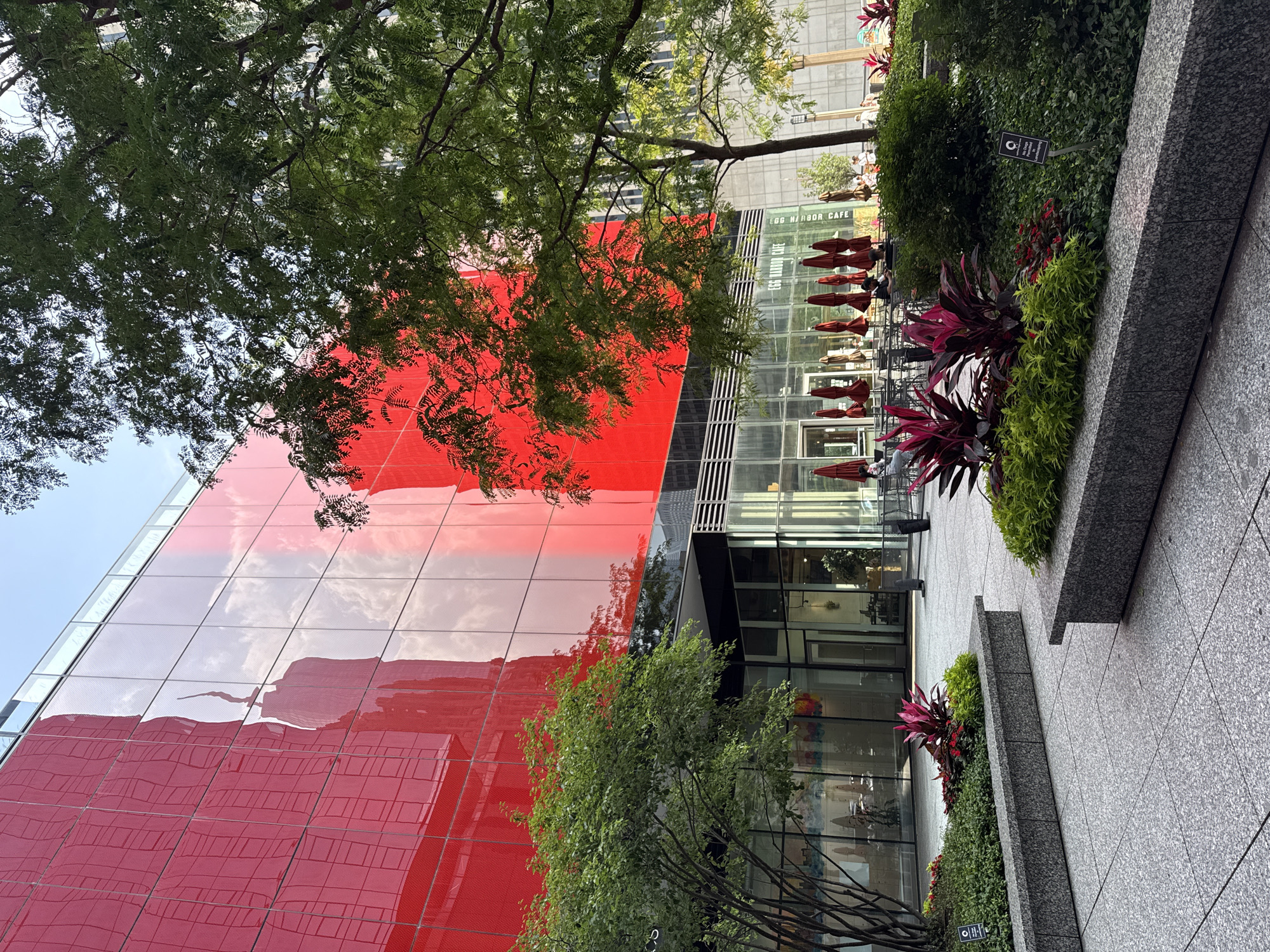 This image depicts an outdoor seating area in front of a modern building with a bright red facade. The building's glass exterior reflects the sky and surrounding structures. Several trees with green foliage are present, providing shade. In the foreground, there are landscaped planters with vibrant red and green plants. A paved walkway leads to the building entrance, where outdoor tables and chairs are set up, some with red umbrellas. People are visible seated at some of the tables, indicating the presence of a cafe or restaurant. A sign in the planter reads "No Dogs Permitted" and has a circle with a line through it, suggesting it's a no-dog zone. The overall scene suggests an urban environment, likely a plaza or courtyard designed for public enjoyment and dining.