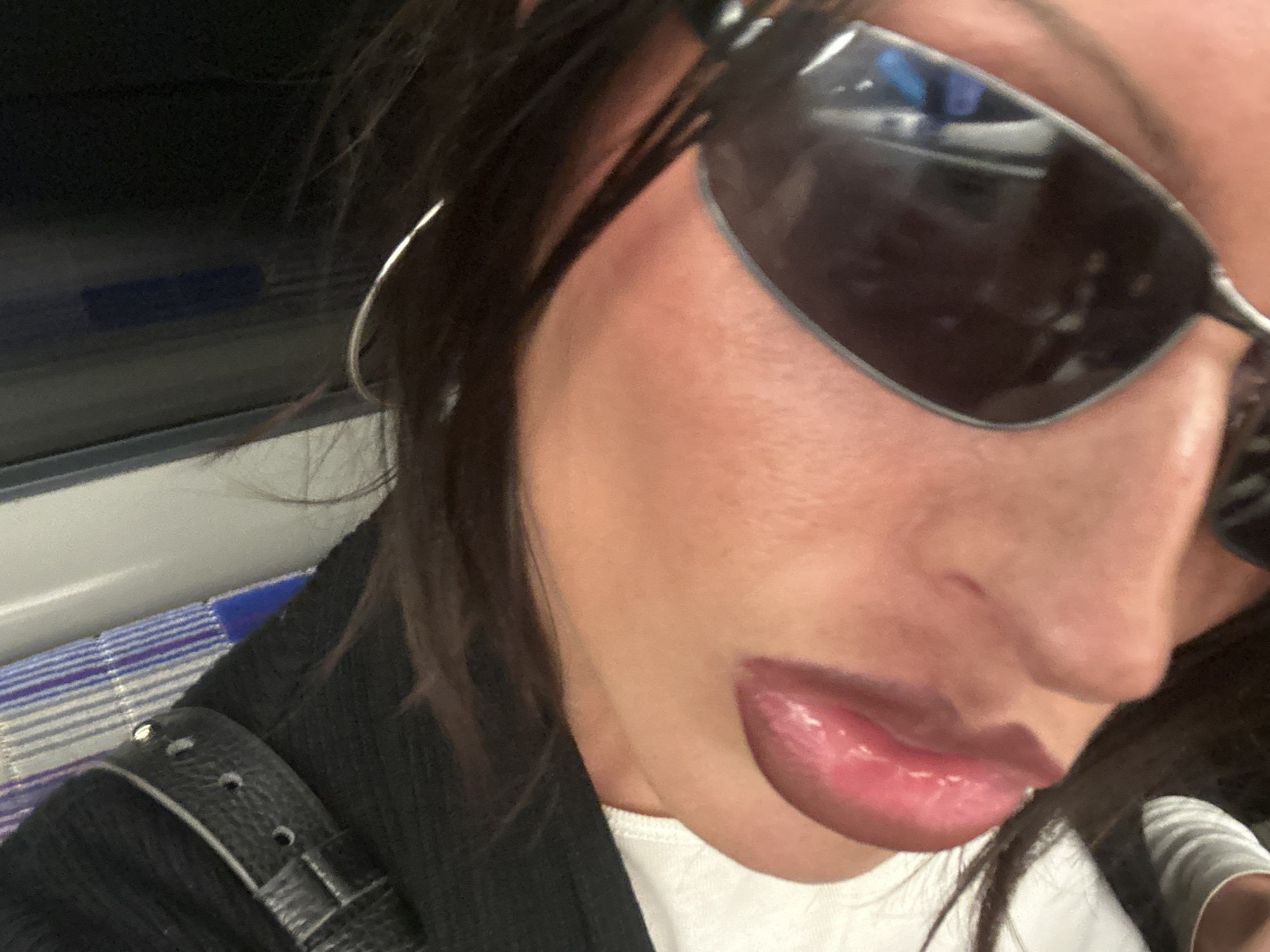 The image presents a close-up, partial view of a person, likely female, captured within a public transport setting in London, United Kingdom. The subject's right side of the face is visible, largely obscured by dark, reflective sunglasses, possibly an aviator style, which show reflections of the interior environment. Dark hair frames her face, and a silver hoop earring is seen on her right ear.
She is dressed in a dark outer garment, which appears to be a jacket or top, featuring a textured strap or buckle detail visible over her left shoulder. Beneath this, a plain white t-shirt is worn. Her lips are full and appear glossy.
The background and surrounding elements strongly indicate a public transport vehicle, such as a train or bus. A distinct blue and white striped pattern, characteristic of seating found in London's public transport system, is visible behind her shoulder. The lighting suggests an indoor setting, and no specific time of day or weather conditions can be determined from the image. No visible text is present. The person appears to be posing casually for a close-up photograph or selfie.