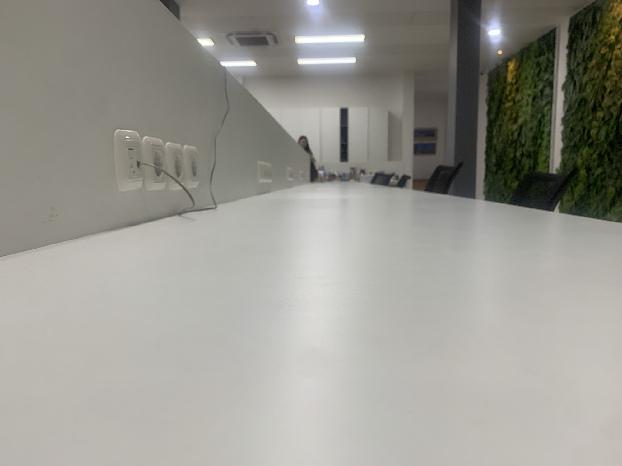 This is a low-angle, wide-shot photograph of an office interior, likely a meeting room or collaborative workspace. The foreground is dominated by a large, white, flat surface, possibly a table, which takes up the majority of the frame and is slightly out of focus.
Along the left edge of the image, a light gray wall is visible. Embedded in this wall are four electrical outlets, three of which are clearly visible. One of these outlets has a black cable plugged into it. A thin, black cable extends from this outlet and runs along the wall.
In the mid-ground, a person is partially visible behind a partition or divider, with their head and shoulders showing. They appear to be looking towards the left side of the frame. Further into the room, several black office chairs are arranged around what is presumably the table.
The background features a modern office environment with a high ceiling. Two long, rectangular fluorescent light fixtures are prominent on the ceiling, emitting a bright, cool light. In the far background, on the right side of the image, is a striking vertical garden wall, covered in lush green foliage, providing a natural element to the otherwise sterile office design. Above the garden wall, another lighting fixture is visible, emitting a warm, yellow light. White cabinetry with dark accents is also present in the background.
The overall impression is a clean, contemporary, and functional office space. The photograph does not provide any specific visual cues that would place it in Palembang, Indonesia.