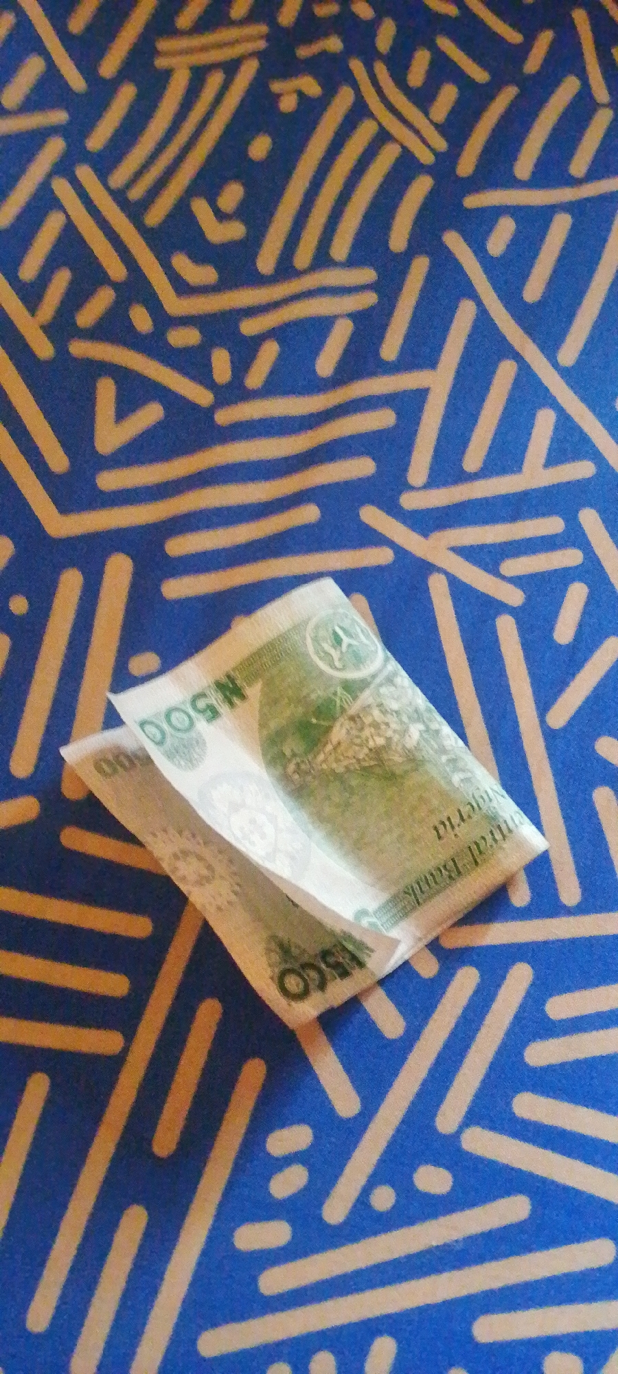 A close-up, slightly overhead shot captures a portion of a Nigerian 500 Naira banknote folded and placed on a blue surface with a yellow abstract pattern. The banknote, primarily green and white, is partially visible, showing segments of its design. The numeral "500" is clearly visible, as are the words "Central Bank of Nigeria" in smaller print on one edge of the bill. The background is a vibrant blue fabric or tablecloth adorned with a repeating pattern of irregular yellow lines and dots, resembling a stylized, scattered design. The lighting suggests an indoor setting, likely during the daytime, with no strong shadows indicating a specific time. The focus is on the banknote and its immediate surroundings, with no people or animals present. The overall impression is a simple, still-life composition focused on currency and pattern.