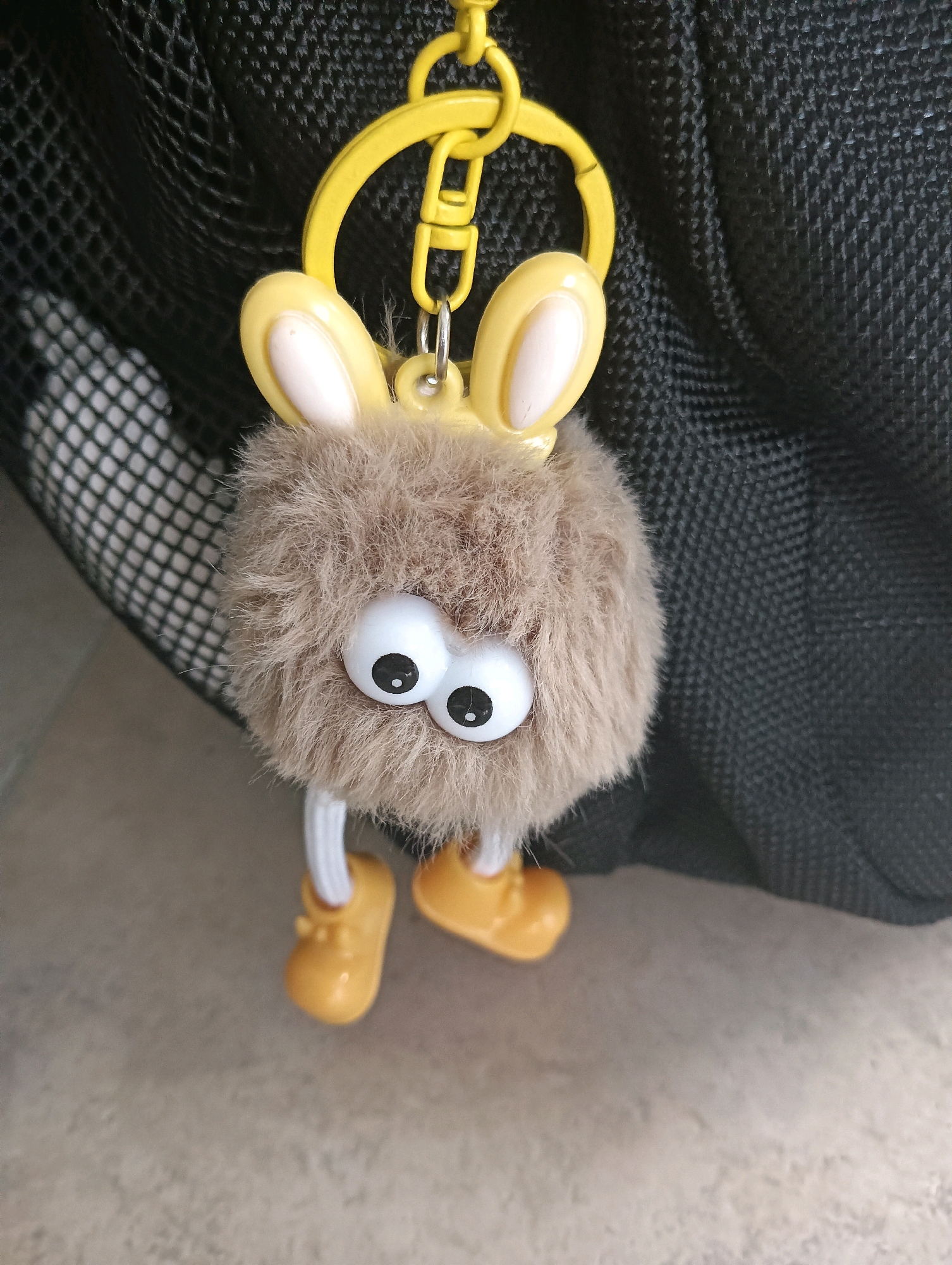 The image shows a close-up of a fluffy, brown, toy keychain with large, googly eyes, yellow bunny ears, and small yellow shoes. The keychain is hanging from a yellow clip attached to a black mesh surface, which appears to be part of a bag. The background is blurred, showing a neutral-colored floor. There is no indication of the location being Tomohon, Indonesia, or any specific city. The scene is static and focuses solely on the keychain.
