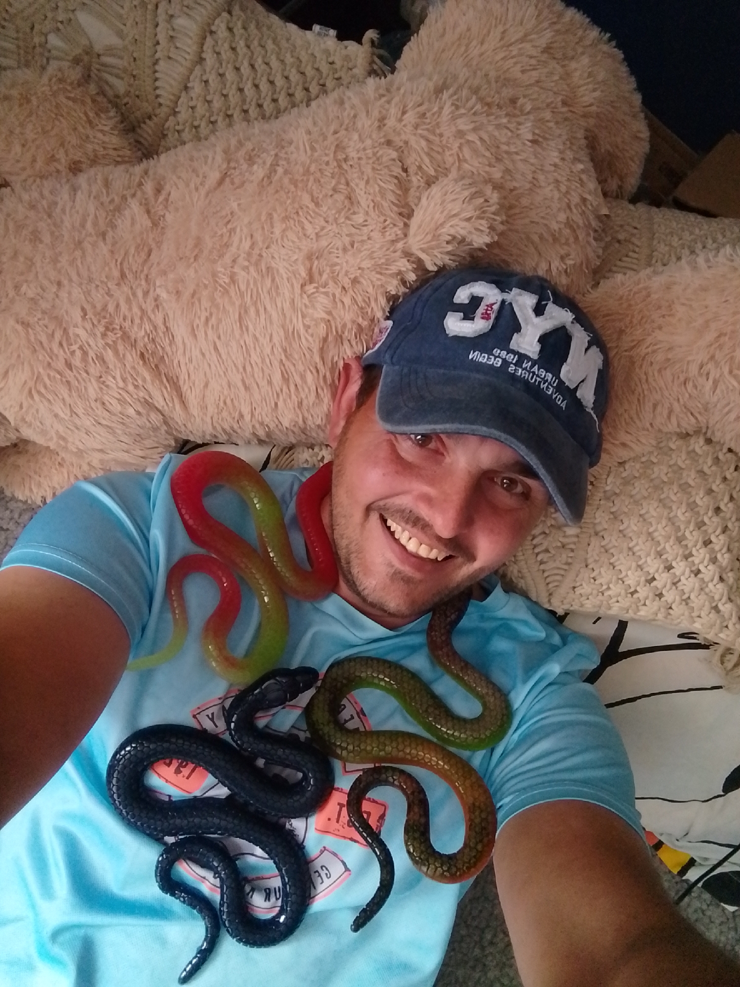A man with a blue cap and a light blue t-shirt is lying on a beige textured blanket and a fluffy beige blanket. He is smiling and has several toy snakes draped around his body. One snake is red and green, another is dark brown, and another is green and brown. The scene appears to be indoors. The location of Wedel, Germany is not ascertainable from the image.