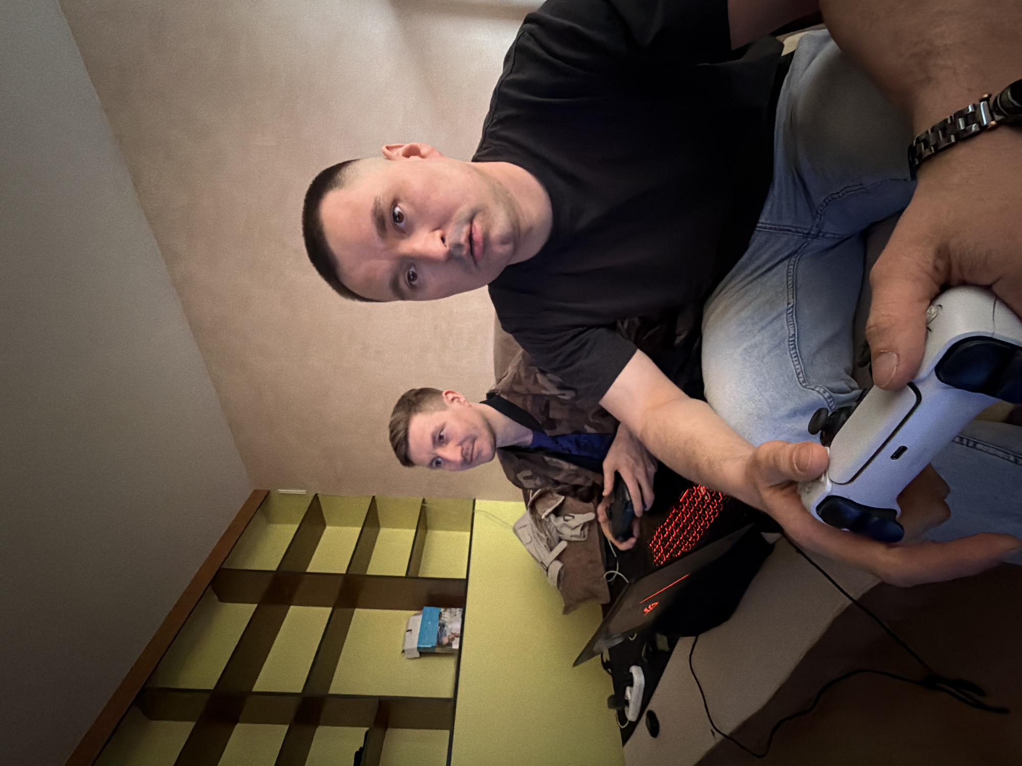 The photograph captures two individuals in what appears to be an indoor environment. The primary subject, a man with a closely shaved head, is wearing a black t-shirt and light-wash jeans. He is holding a white Playstation 5 controller. He has a watch on his left wrist. Below him, another man, wearing a camouflage jacket, is using a computer and a mouse. An illuminated red gaming keyboard is visible on a laptop. A bookshelf with multiple compartments can be seen in the background, along with various objects including a bag. The photograph is taken at an angled perspective. The location is not evident from the image. There is no indication of Izhevsk, Russia, or its urban environment.