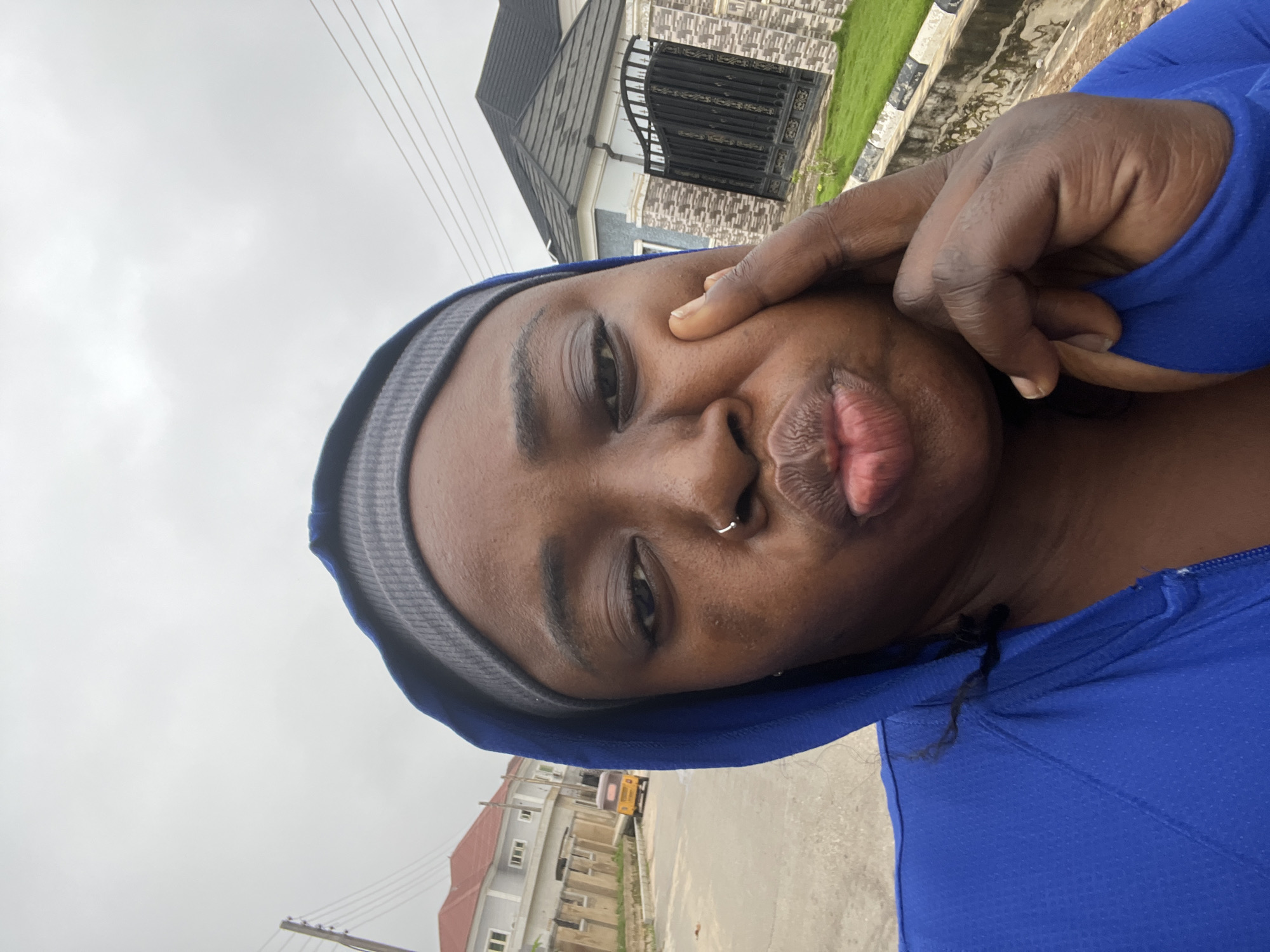 A selfie taken outdoors in what appears to be a residential area of Abuja, Nigeria, shows a dark-skinned woman wearing a blue hooded jacket and a grey headband. She has a nose ring and is puckering her lips, with her right hand touching her cheek. In the background, a yellow auto-rickshaw is visible on a street, along with modern houses and a black metal gate. The sky is overcast.