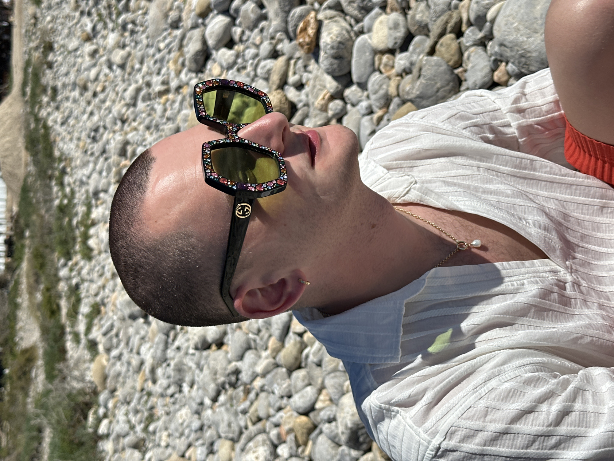 A light-skinned male with a shaved head is lying on a rocky beach. He is wearing bedazzled sunglasses, a white textured shirt, a gold chain necklace with a pearl pendant, and a gold hoop earring. His mouth is slightly open and he is gazing upwards, towards the left of the frame. The location appears to be a beach near Sant Josep de sa Talaia, Spain, evidenced by the presence of small, smooth, grey-colored rocks. The weather is sunny, casting shadows on the white shirt. There are green plants visible in the blurry background on the left.