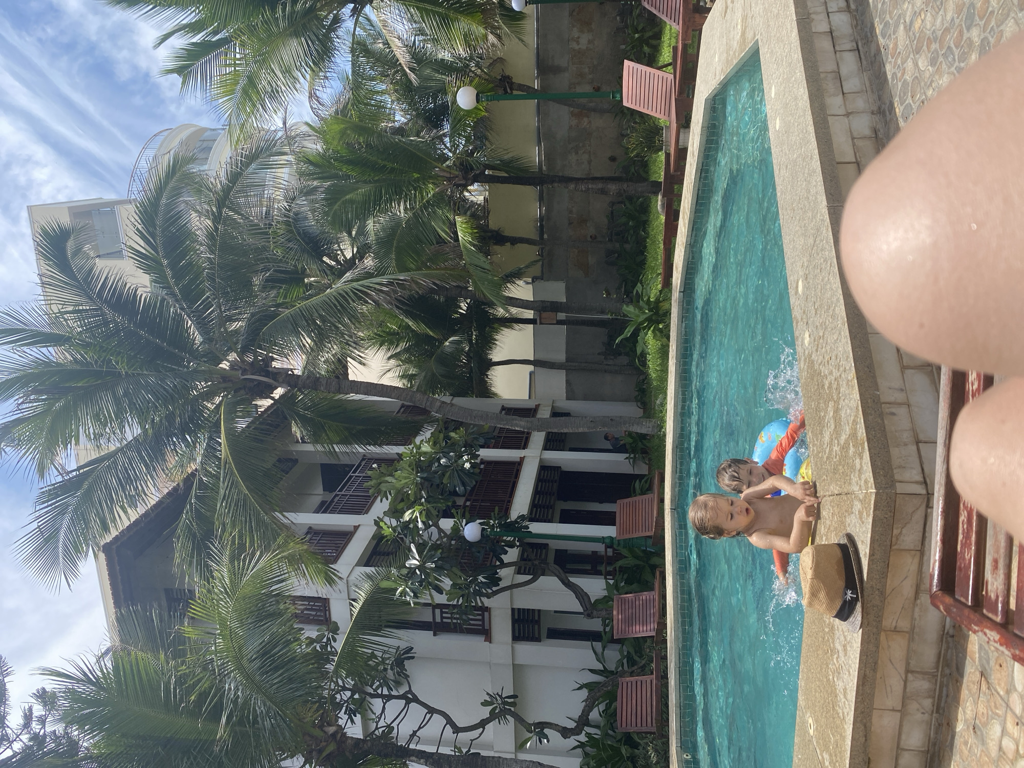 In Phu Thuy Ward, Vietnam, a family enjoys a day by the pool, surrounded by lush palm trees and modern architecture. The bright, sunny day illuminates a refreshing swimming pool, where two children, a girl and a boy, splash and play. The girl, with blonde hair, is reaching into the water, while the boy, with brown hair, is wearing an orange and blue inflatable ring, his face partially obscured by water. Nearby, a straw hat with a black band rests on the pool's edge. In the background, a multi-story white building with brown balconies stands tall, providing shade and accommodation for guests. Wooden lounge chairs are strategically placed around the pool, inviting relaxation. The overall scene captures a moment of joy and leisure in a tropical setting.