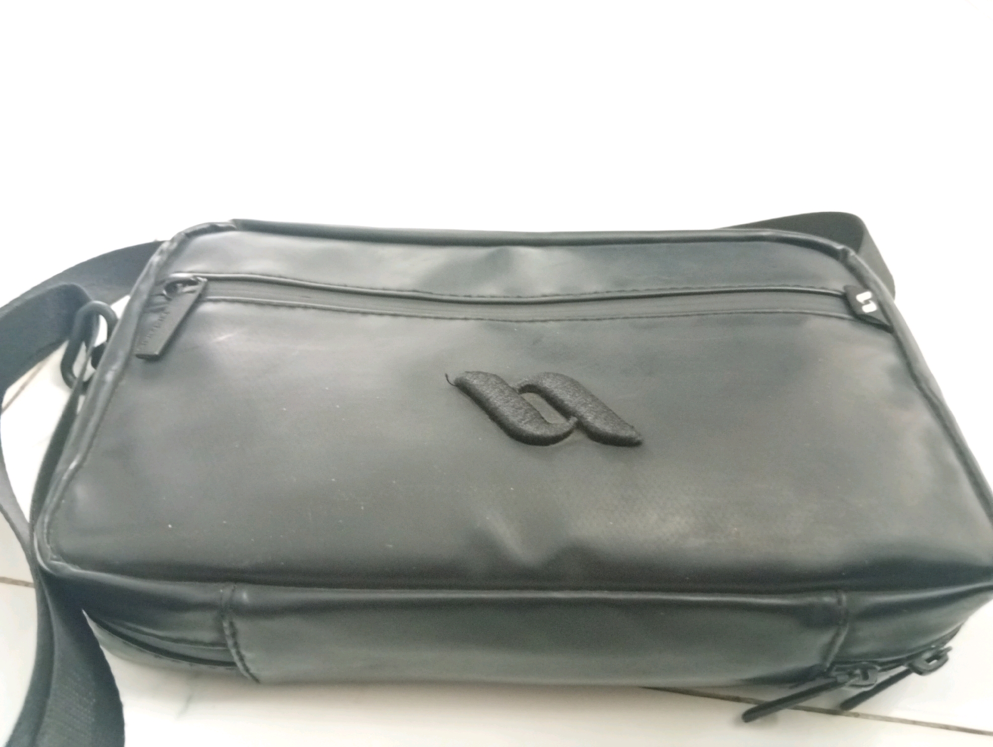 A black crossbody bag with a prominent dark gray embossed logo is laid on a white tiled surface. The bag has a zippered compartment on the front, and zippers are visible on the right side and bottom. The strap is black and appears to be made of nylon. The lighting is bright, suggesting an indoor environment with a white background, possibly a home or a retail space in Samarinda, Indonesia.