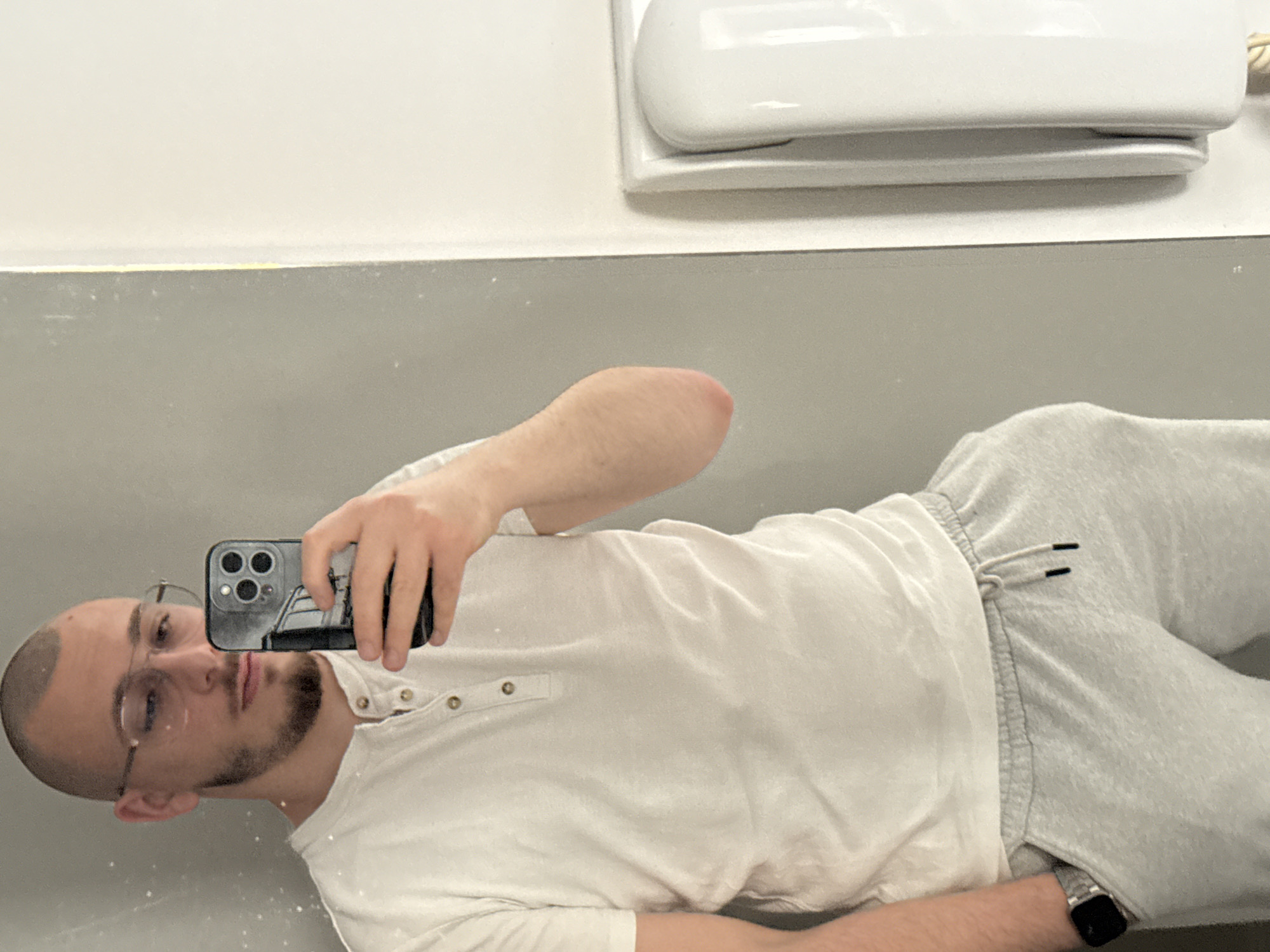 A young adult male, likely in his 20s or early 30s, is depicted in a mirror selfie taken within an interior space in Stara Zagora, Bulgaria. The subject has a shaved head, a short beard, and wears thin-framed glasses. He is dressed in a white, short-sleeved Henley shirt with three visible light brown buttons, and light grey drawstring sweatpants. His left hand is partially inserted into the left pocket of his sweatpants, revealing a silver-banded smartwatch on his left wrist. His right hand holds a dark-colored smartphone, featuring a triple camera array, which he uses to capture his reflection. The phone's screen partially obscures his face.
The background visible in the mirror consists of a light grey wall to the left and a white wall to the right. A thin vertical yellow line marks the seam where the grey and white surfaces meet. On the far right, a white, vertically oriented wall-mounted dispenser, consistent with a soap or paper towel dispenser, is partially visible against the white wall. The environment suggests a public or commercial restroom or a similar utility space. The lighting is bright and even.