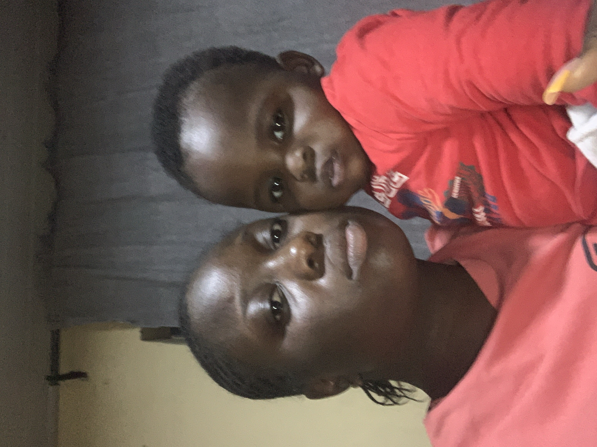 In a dimly lit indoor setting, a woman and a baby are captured in a close-up selfie. The woman, positioned on the left, is wearing a pink shirt and her hair is styled in braids. The baby, on the right, is dressed in a red shirt and is being held by the woman. The background consists of a plain wall and a gray curtain. The lighting is soft, with a noticeable sheen on the subjects' skin, suggesting indoor artificial lighting. The overall mood is intimate, likely a candid moment captured by the woman herself.