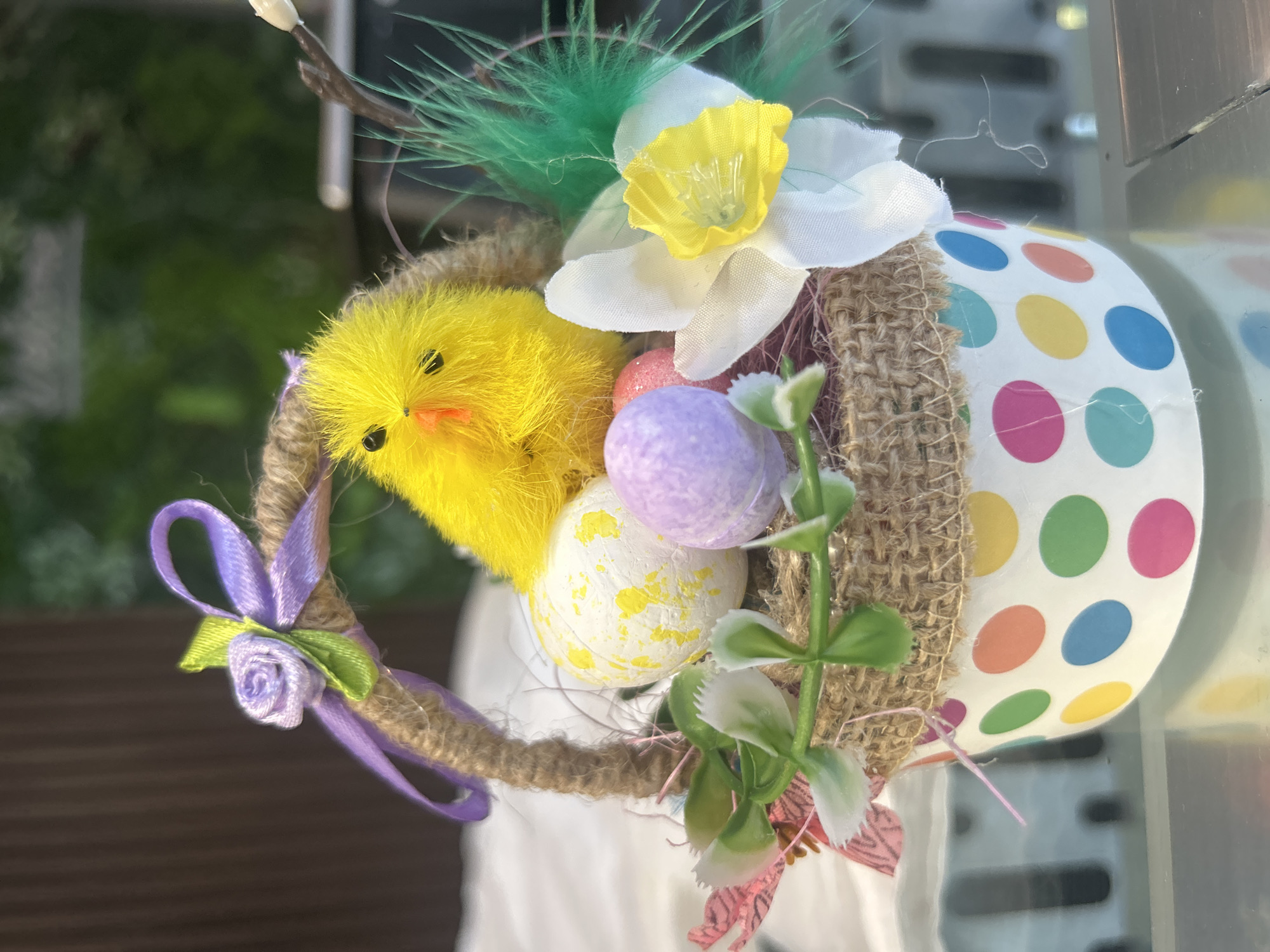 An Easter basket arrangement is presented, featuring a fuzzy yellow chick adorned with orange beak and black eyes, placed amongst decorative Easter eggs. One egg is white with yellow speckles, another is a solid pale purple, and a third is pink. The basket itself is constructed from burlap, with a handle wrapped in twine, and decorated with a purple ribbon and a small fabric rose. Green artificial leaves and a white daffodil with a yellow center are also incorporated into the arrangement. The basket is contained within a decorative paper cup adorned with colorful polka dots. The background includes a blurry brown surface and green foliage, suggesting an indoor setting with an outdoor view. The image was taken in Varna, Bulgaria.