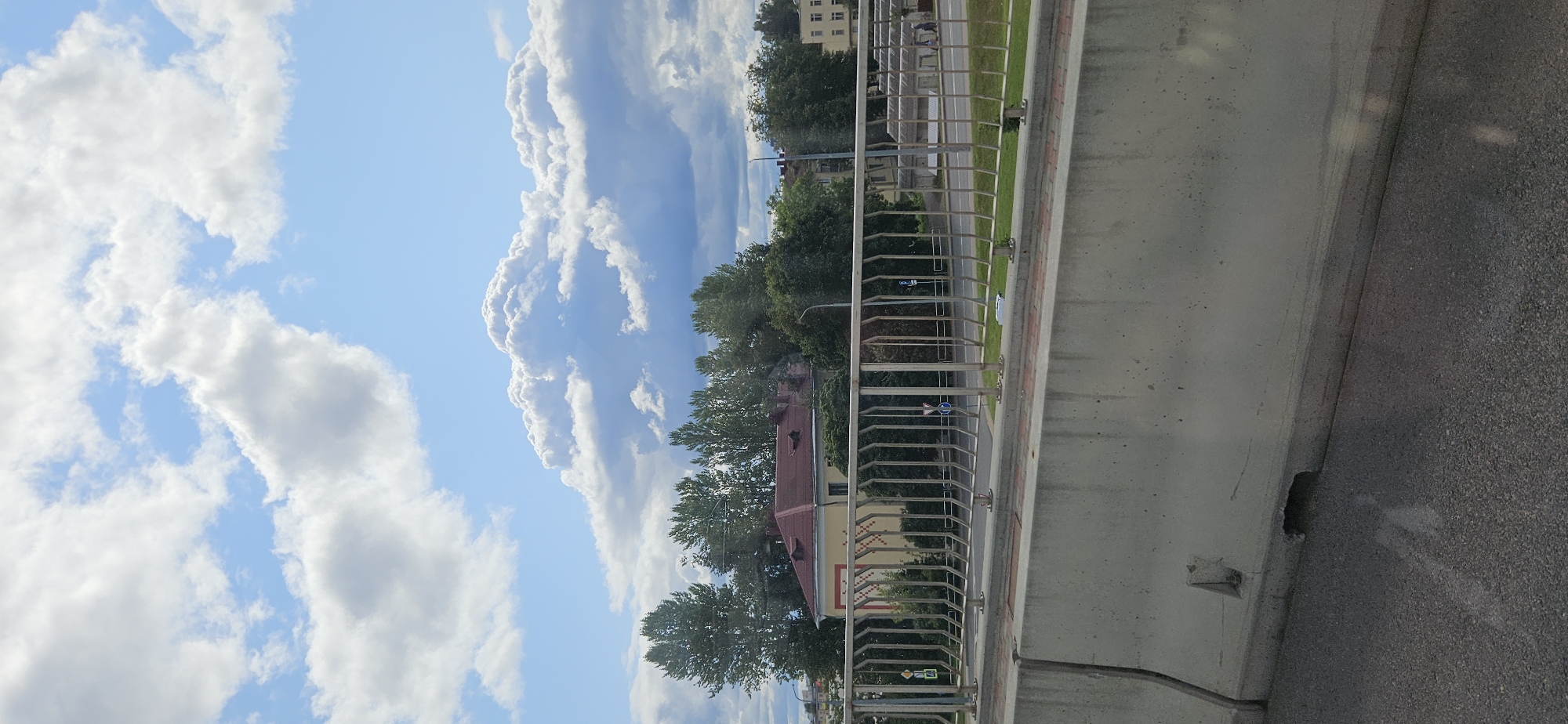 The image captures a view from a raised position, likely a bridge or overpass, overlooking a street and some buildings in the city of Vitsebsk, Belarus. In the foreground, a grey concrete barrier runs horizontally across the frame, with a metal railing positioned above it. Beyond the railing, a street is visible, with a grassy area bordering it and a row of trees in the mid-ground. A building with a red roof and yellow walls, featuring decorative patterns and dormer windows, is prominent among the trees. Further in the distance, other buildings and structures can be seen, including what appear to be garages or sheds with grey roofs. The sky above is a bright blue, dotted with numerous white, fluffy clouds, suggesting fair weather. The overall scene depicts an urban environment with a mix of infrastructure and greenery under a dynamic sky.