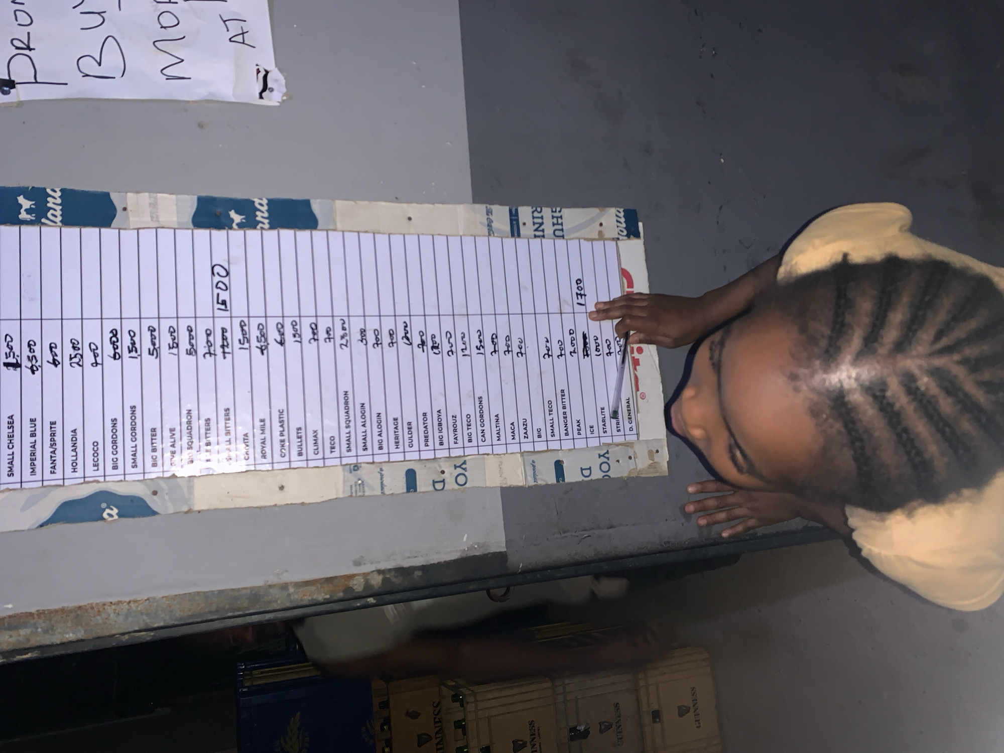 A young child with braided hair is looking at a price list for various beverages. The price list, affixed to a wooden backing, includes brands like "Small Chelsea," "Imperial Blue," "Fanta/Sprite," and "Hollandia." Prices, measured in local currency, are listed next to each item. The setting appears to be a store or business environment, possibly located in New Stadium, Nigeria, as suggested by the presence of Guinness crates in the background. A sign with the text “Promo Buy More At” appears at the top left of the frame.