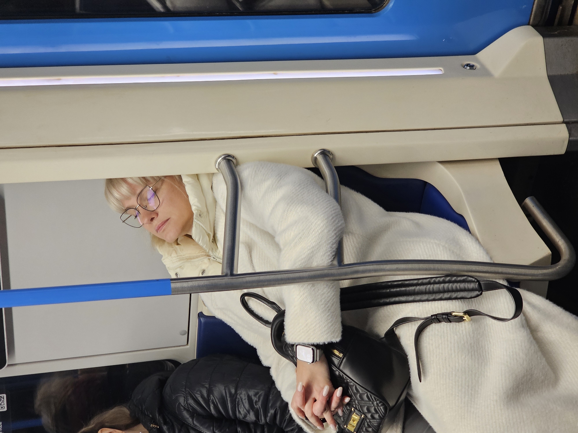 The image displays the interior of a public transport vehicle, likely a metro train in Moscow, Russia, characterized by its clean, modern design featuring light cream-colored panels and contrasting blue accents, particularly on the overhead rack and seat upholstery.
The main subject is a blonde woman with glasses, seated comfortably. She is wearing a cream-colored, textured coat over a lighter, possibly quilted, jacket. Her head is tilted slightly, and she appears to be resting or looking downwards, perhaps at a phone or book not visible in the frame. On her left wrist, she wears a silver-toned smartwatch with a lit screen. Her hands, featuring light-colored manicured nails, are clasped over a black quilted handbag with a prominent gold-colored buckle or logo on its strap.
Metal handrails are visible in the foreground, indicating passenger support features within the vehicle. A bright, strip-like light fixture runs along the upper part of the interior paneling. Below the main subject, to the left, parts of another person's head and shoulder are visible, clad in a black puffer jacket, suggesting another passenger seated nearby.
The scene conveys a quiet moment during transit, likely a daily commute. The lighting is uniform and artificial, typical of an indoor environment like a subway car. A very small, partially obscured QR code and the word "FREE" are faintly visible on a white panel to the lower left, behind the blue handrail.