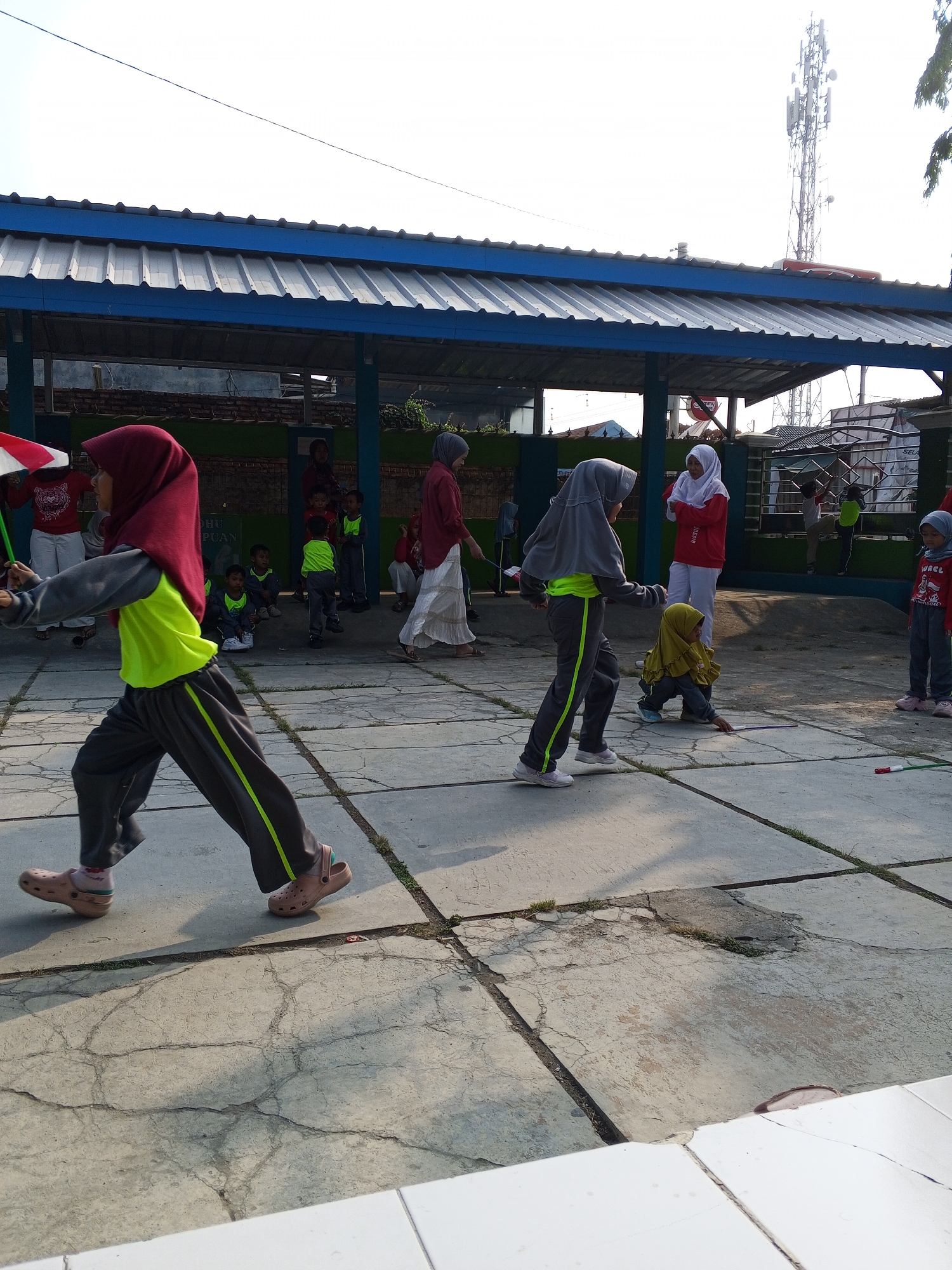 In the city of Gebang, Indonesia, a group of children are engaged in an outdoor activity on a paved area under a covered structure. The activity appears to be some form of relay race or game involving flags. One child in the foreground, wearing a red hijab and a neon yellow and grey tracksuit, is mid-stride, holding a green and white flag. Several other children, some in similar tracksuits and head coverings, are dispersed around the paved area, some observing and others participating. In the background, there is a brick wall and a cell tower. The ground is made of large concrete slabs with visible cracks and patches of grass growing through them. The lighting suggests it is daytime, with clear skies and shadows cast by the structure and the children.