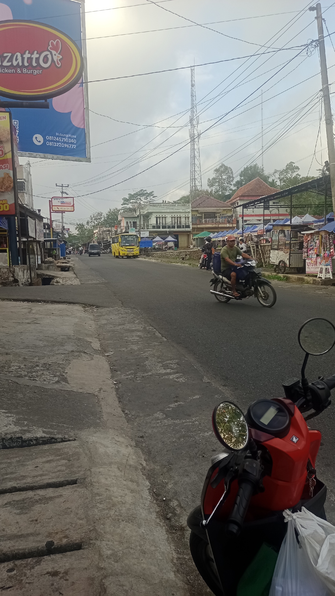The image depicts a street scene in Pakemitan, Indonesia. The foreground features the front portion of a red scooter with a side mirror reflecting trees. The road itself is paved and shows signs of wear and tear.
Further down the road, a yellow bus is visible, along with several motorcycles and scooters carrying passengers. On the sides of the road, there are buildings with shops and market stalls. One prominent sign advertises "Chitato Chicken & Burger." In the background, telecommunication towers rise above the buildings, and power lines crisscross the sky, which is overcast. The overall impression is of a busy, commercial street in a semi-urban or rural Indonesian setting.