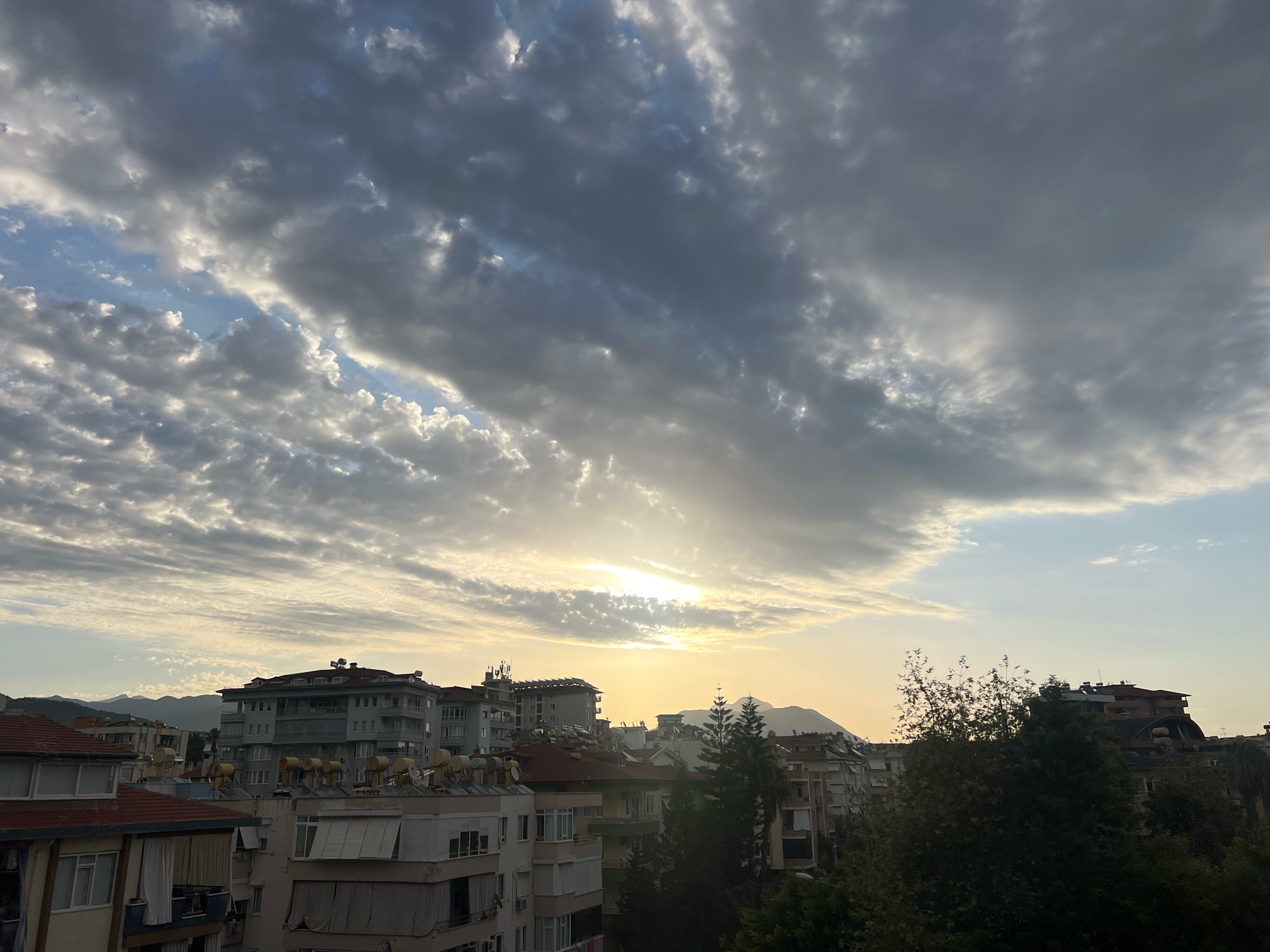 The image shows a cityscape in Alanya, Turkey, under a cloudy sky with the sun peeking through. The foreground features the tops of several multi-story apartment buildings with red-tiled roofs and solar water heaters visible. Some buildings have visible air conditioning units and satellite dishes. Evergreen trees are scattered throughout the urban landscape. In the background, rolling hills or mountains are visible under a hazy sky. The lighting suggests it is either sunrise or sunset. There are no people or animals visible in the scene.