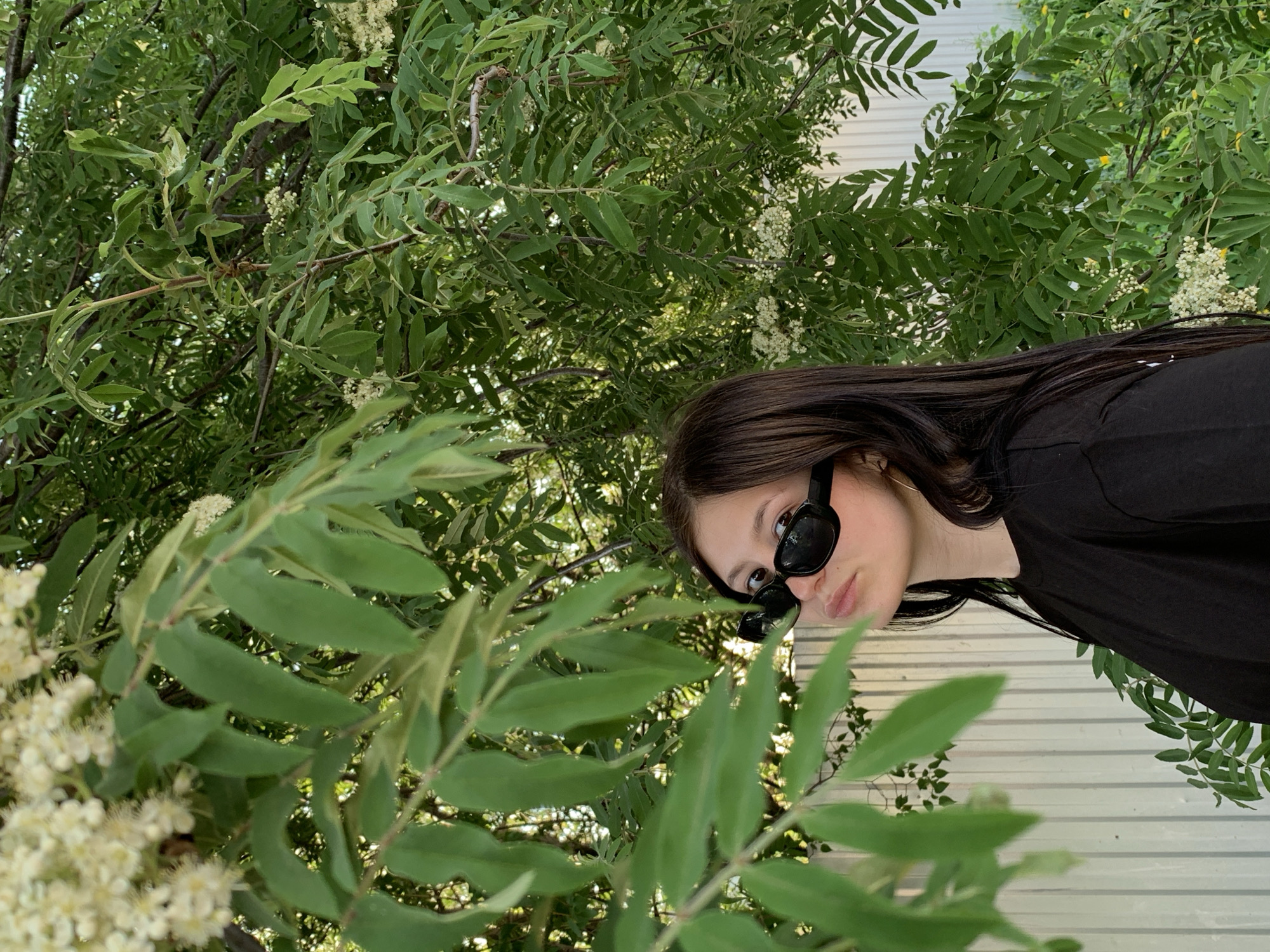 A medium shot features a young woman with long, dark hair and sunglasses. She is wearing a black shirt and positioned to the right, with her face tilted towards the viewer. The background and foreground are filled with lush green foliage and small white flowers. A light grey wall is visible behind the foliage and woman, indicating a possible building or fence structure. The location is given as Фоки, Russia, but no elements in the image definitively confirm this.