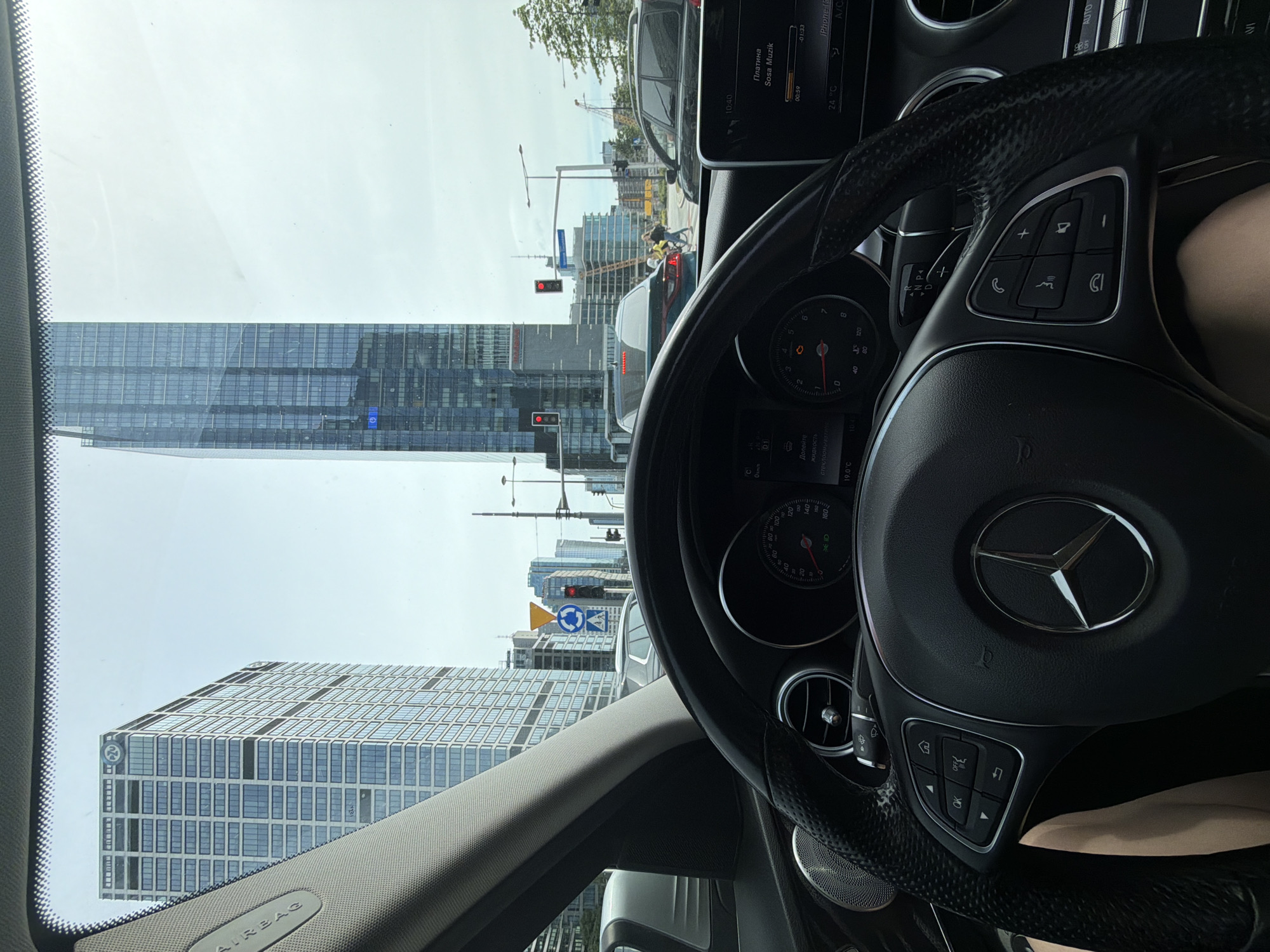 The image shows the interior of a car, specifically a Mercedes-Benz, with the steering wheel prominently displayed. Through the windshield, a cityscape is visible. There is a tall, modern skyscraper with a glass facade and another building with a similar design visible. The traffic light is red. The OCR on the infotainment screen displays information in Russian and English, including the time (10:40), the song playing (Sosa Musik), and the temperature (24°C). There is also a circular blue road sign visible on the street. The location is likely in a city with modern architecture, possibly in Eastern Europe or a region where Russian is spoken.