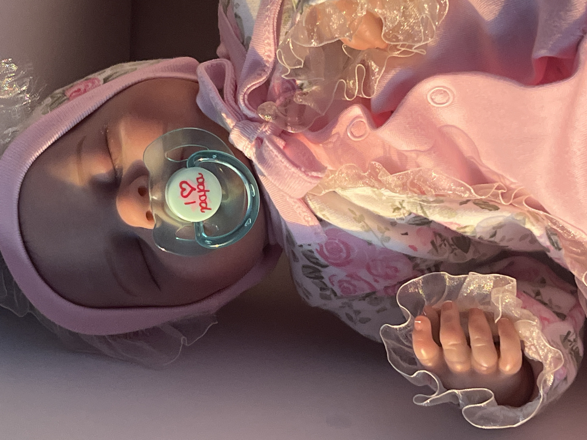 The image shows a close-up, overhead shot of a newborn doll lying down. The doll is wearing a pink bonnet and a pink ruffled outfit with a floral pattern. A pacifier with the word "papa" and a heart symbol is in the doll's mouth. The doll's hand, with visible fingers, is peeking out from under the ruffled outfit. The setting appears to be a box or a bassinet, with a soft, diffused light suggesting indoor lighting. The overall impression is one of tenderness and a focus on the doll's features.