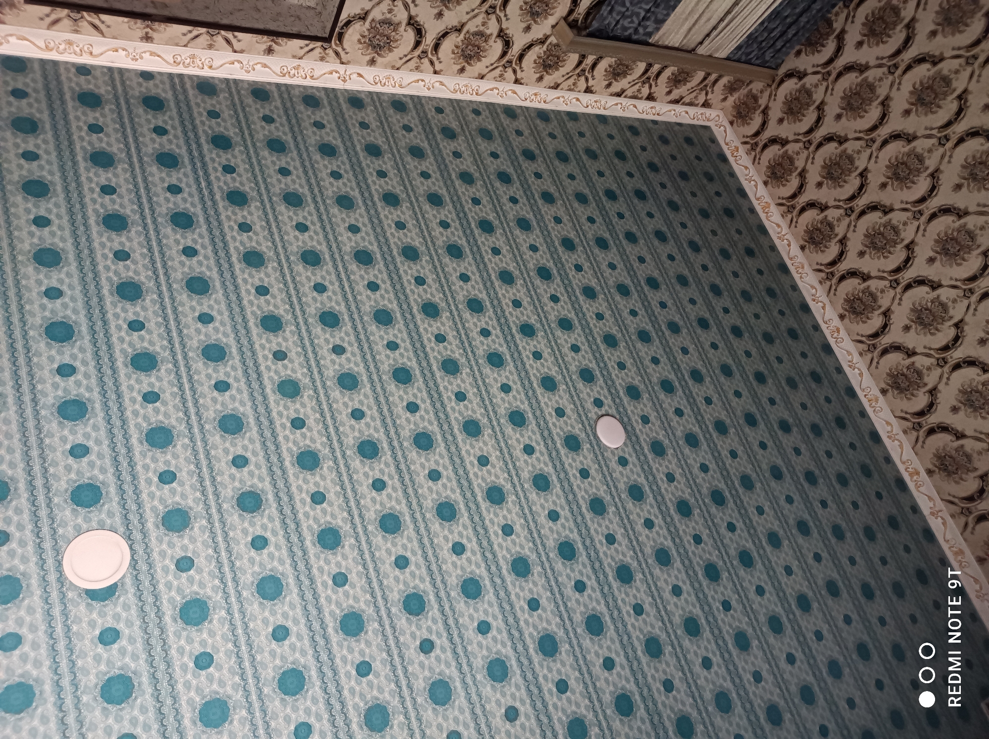 The image shows a patterned ceiling with turquoise circles and decorative borders. Two white circular light fixtures are visible on the ceiling. The ceiling is adorned with horizontal bands of patterned wallpaper, featuring a repeating motif of small and large turquoise circles. A decorative white and gold border runs along the edge of the ceiling, transitioning to a wall covered in a floral patterned wallpaper. The floral pattern on the wall is a light beige with darker brown and black floral designs. The lower right of the image shows a corner where the ceiling meets the wall and possibly another wall or a piece of furniture with striped fabric. The lighting in the image appears to be artificial, casting a slight shadow and highlighting the textures of the materials. The scene depicts an interior space, likely a room. There are no people visible in the image. The overall impression is of a decorated interior with a focus on patterned surfaces.