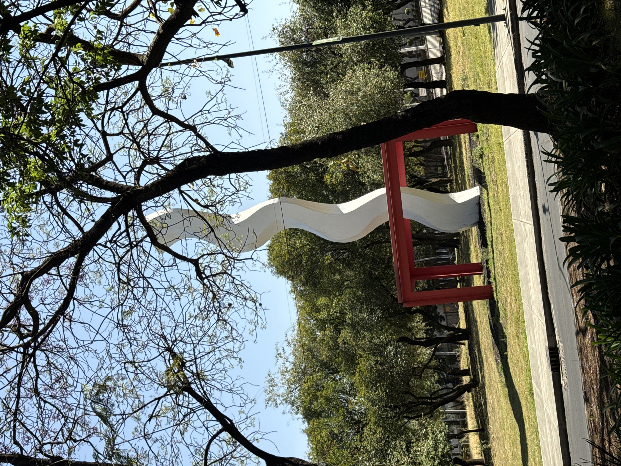 The image captures a park scene in Mexico City, Mexico. The foreground is dominated by trees with a mix of bare branches and emerging leaves, indicating either late winter or early spring. A modern, undulating white sculpture, framed by a red metal structure, stands prominently in the midground. Behind the sculpture, a line of lush green trees forms a natural backdrop, contrasting with the clear blue sky visible through the branches. A concrete sidewalk runs along the edge of the park, bordered by a strip of manicured green grass. In the distance, a glimpse of urban architecture and utility poles suggests the park's location within a city setting. The overall impression is one of a tranquil urban oasis, blending nature with modern art.