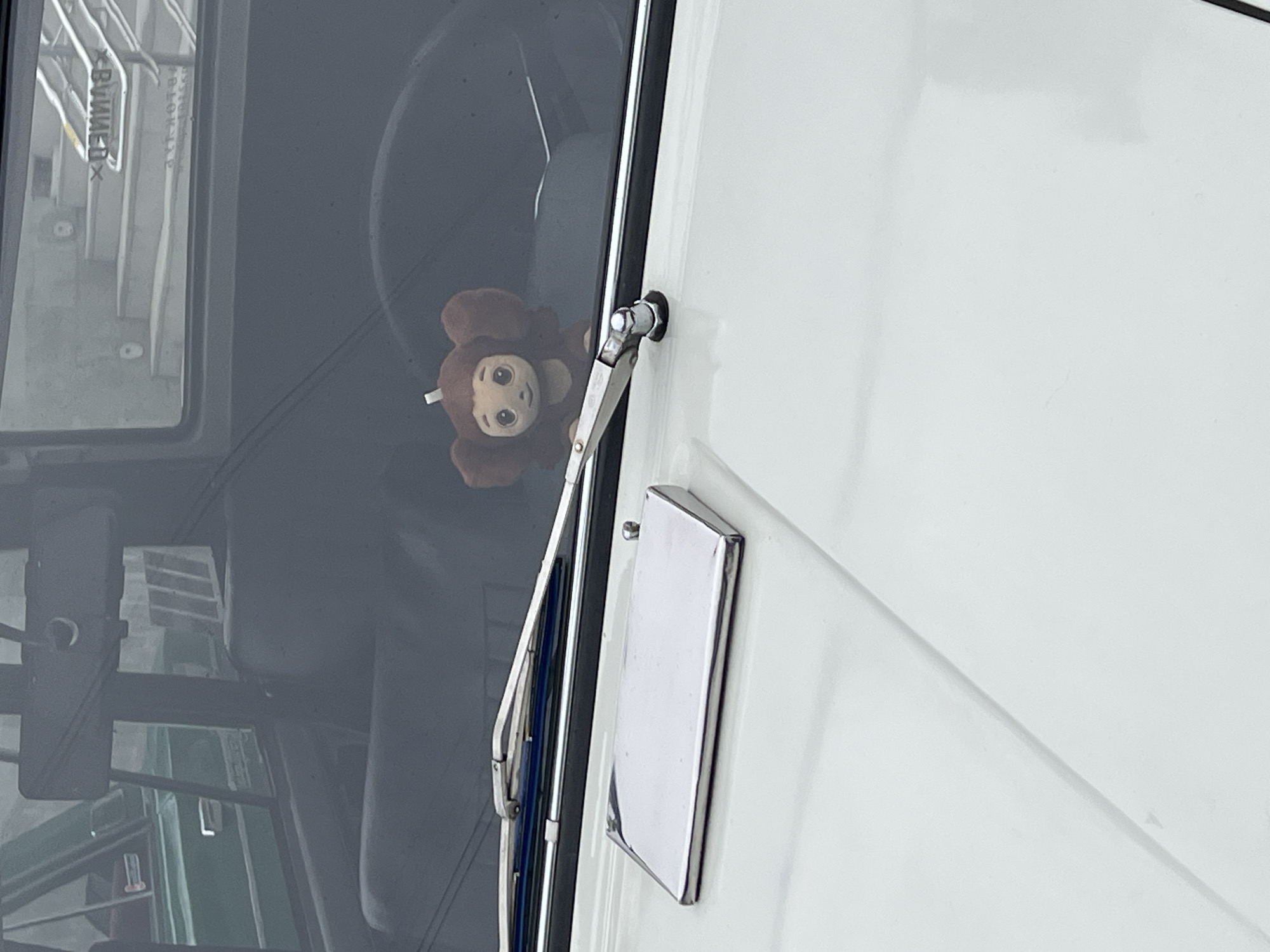The image depicts a Cheburashka plush toy positioned inside the windshield of a vehicle or boat, seemingly in Saint Petersburg, Russia, based on the Cyrillic script visible through the glass. A wiper blade is seen on the exterior of the glass, and a rectangular silver object is attached to a white surface beside the window.