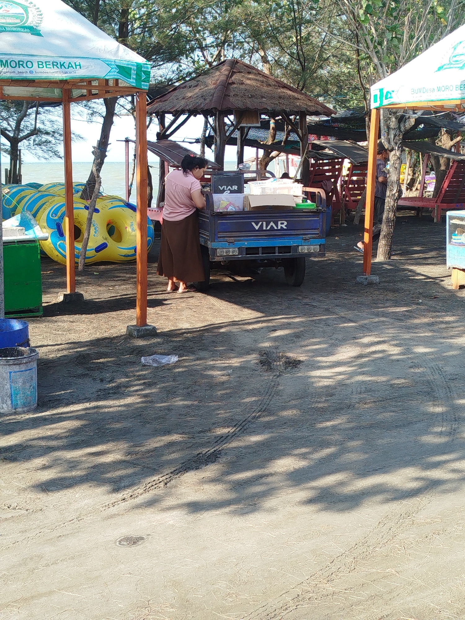 The image captures a scene on the beach in Mororejo, Indonesia. In the foreground, sandy ground is visible with tire tracks and shadows cast from trees overhead. To the left, a green plastic container and a blue bucket are present. In the middle ground, a woman wearing a pink shirt and brown skirt is standing next to a blue truck. The truck has "VIAR" written on its side and appears to be loaded with boxes. Behind the woman and truck, several yellow and blue inflatable tubes are stacked. A small hut with a thatched roof is also visible. In the background, trees and the ocean can be seen. A white tent with text that includes "BUMDes MORO BERKAH" is situated to the left. The overall environment suggests a recreational area at the beach.