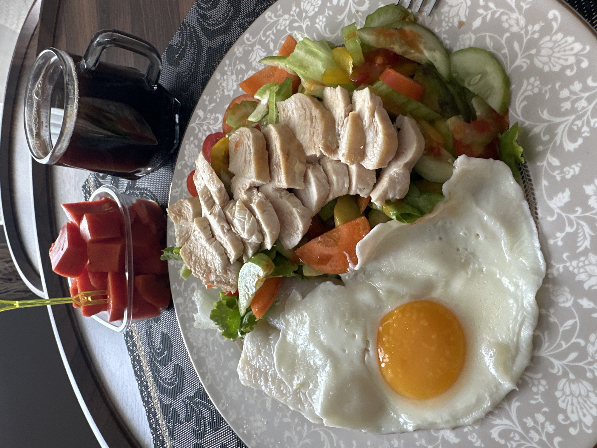 A top-down medium shot shows a plate of food on a table in Pattaya City, Thailand. The plate contains a salad with sliced chicken, cucumbers, tomatoes, and lettuce. Next to the salad is a fried egg. A glass of dark liquid and a small container of cubed red fruit sit to the left of the plate. The table surface is covered with a patterned placemat. A table edge is visible in the lower left corner. The overall lighting is bright and natural.