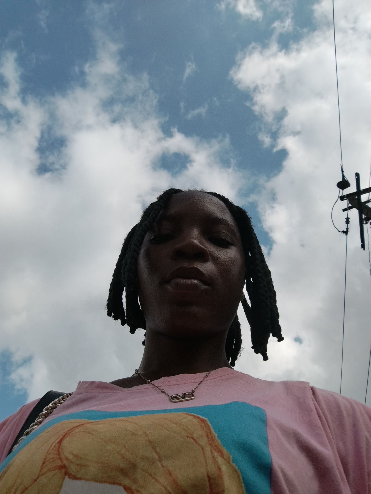 The image shows a low-angle, waist-up shot of a young Black woman with dark skin and black braided hair, outdoors under a cloudy sky, likely in Benin City, Nigeria. She is wearing a pink t-shirt with a graphic print and a silver chain with a pendant. To the right of the frame, a utility pole with wires is visible against the sky. The woman's face is looking directly at the camera. The lighting suggests it is daytime.