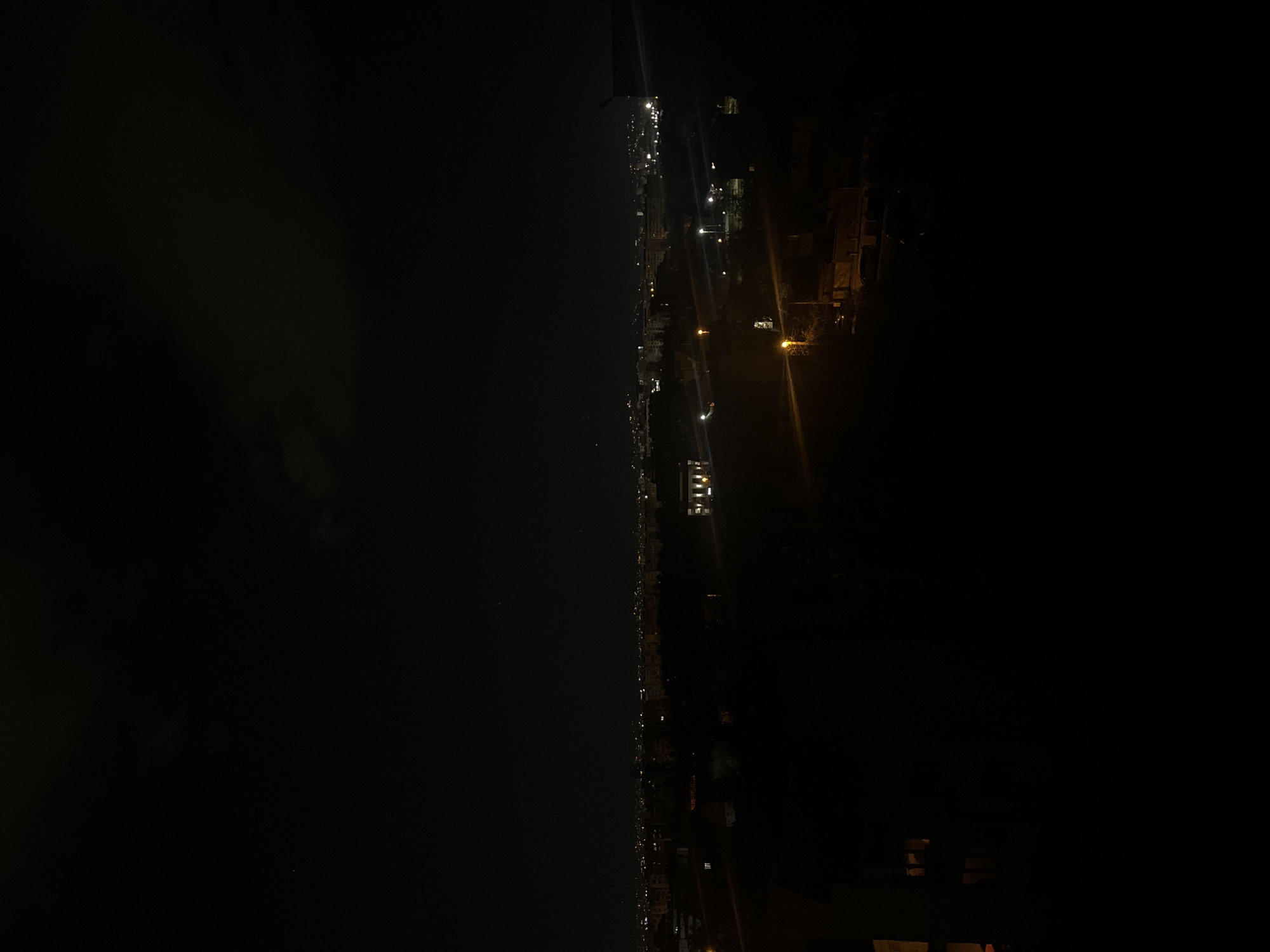 The image shows a night view, likely from an elevated position in Sofia, Bulgaria. A line of lights extends across the center of the frame, suggesting a coastal road or waterfront area. To the left of this line is a dark expanse, possibly the Black Sea or a large body of water. To the right, the scene is mostly dark, but some streetlights and building lights are visible, indicating urban development. Overall, the image conveys a scene of a lit coastline juxtaposed with dark water and urban areas at night.