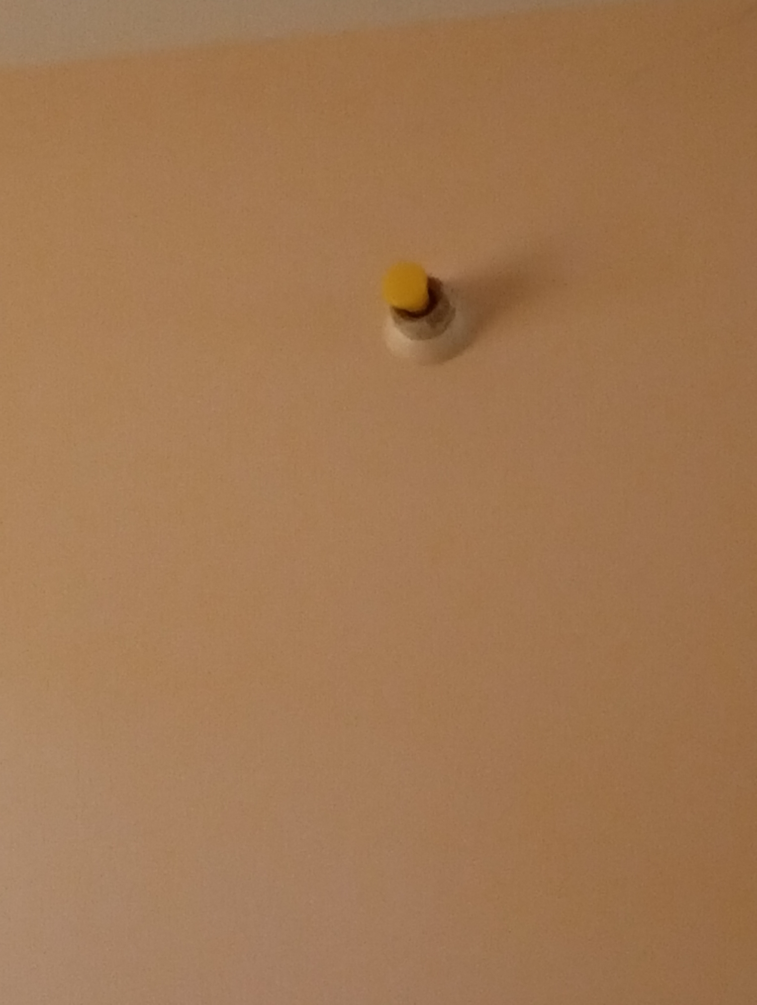 A yellow button is mounted on a white circular fixture on a textured, light orange wall. The fixture appears to be a smoke detector or a similar type of sensor. The background is a plain wall. The image is taken from a low angle looking upwards.